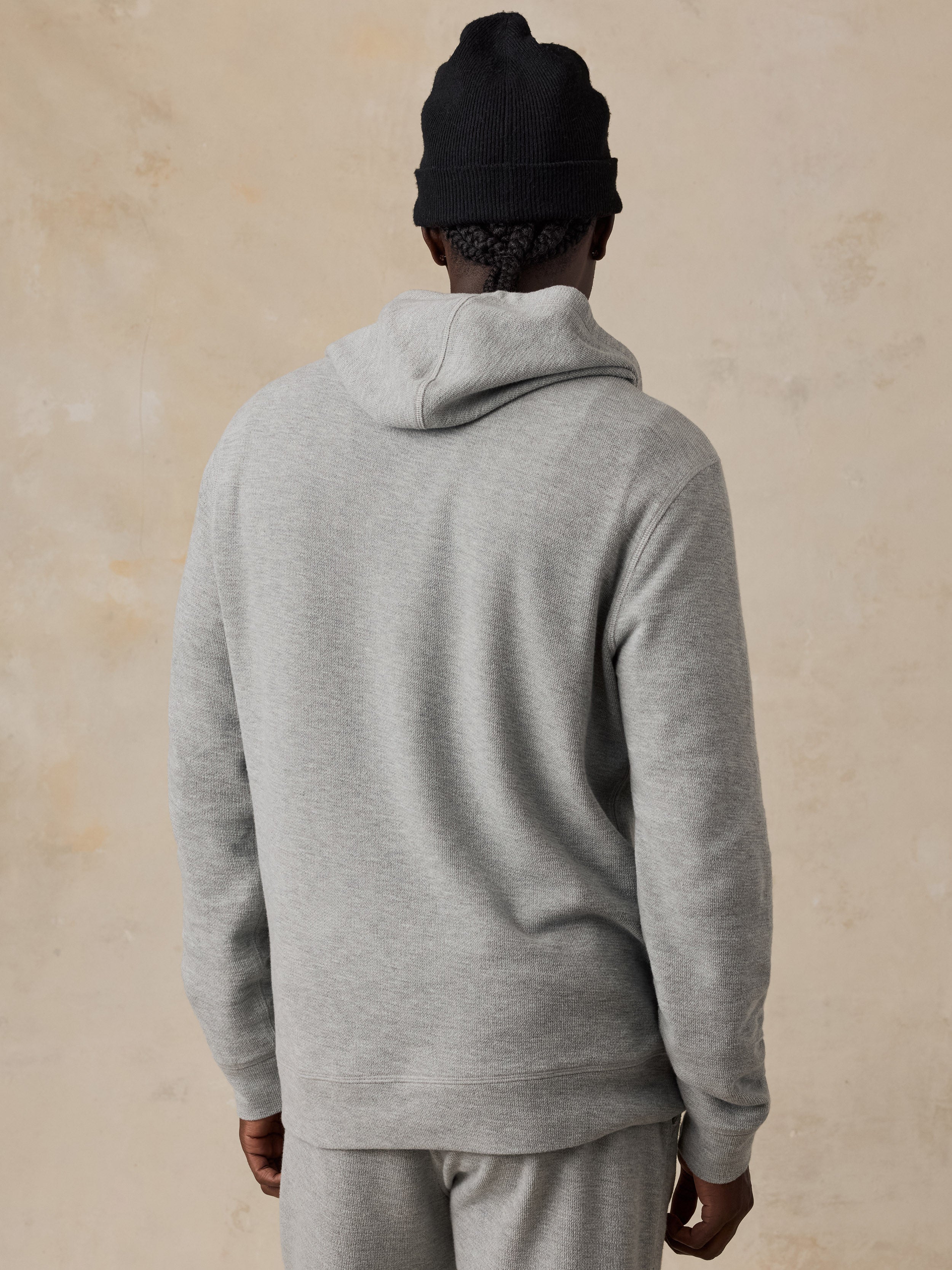 A man shown from the back wearing a light heather gray hooded sweatshirt and matching gray sweatpants, with a black knit beanie, against a warm beige textured wall.