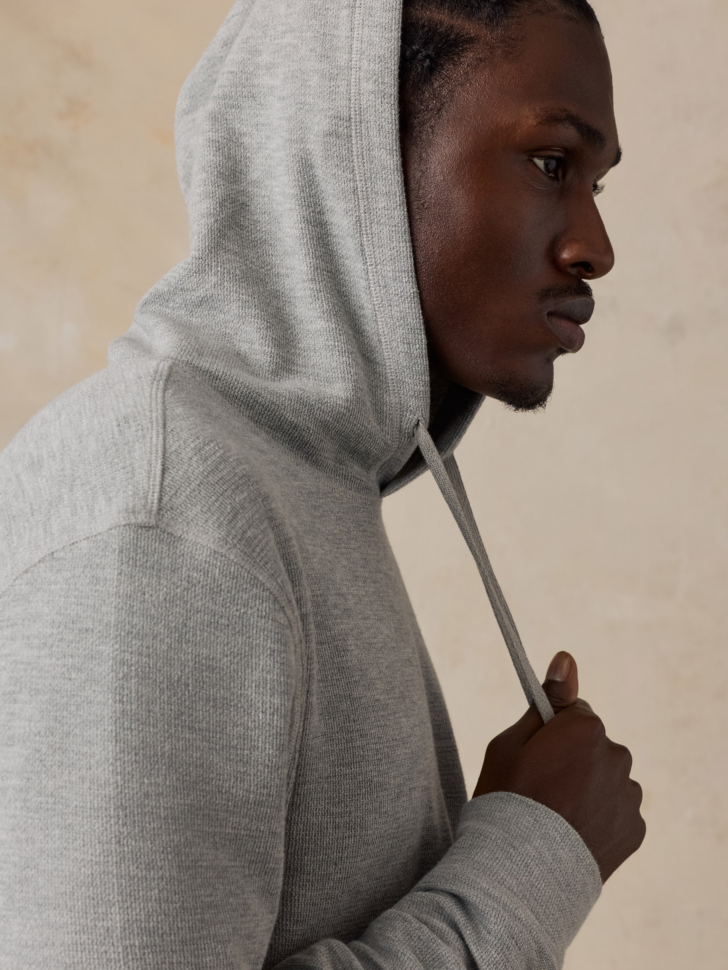 A man in profile wearing a light gray heathered hoodie, holding the drawstring near his chest against a neutral beige background.