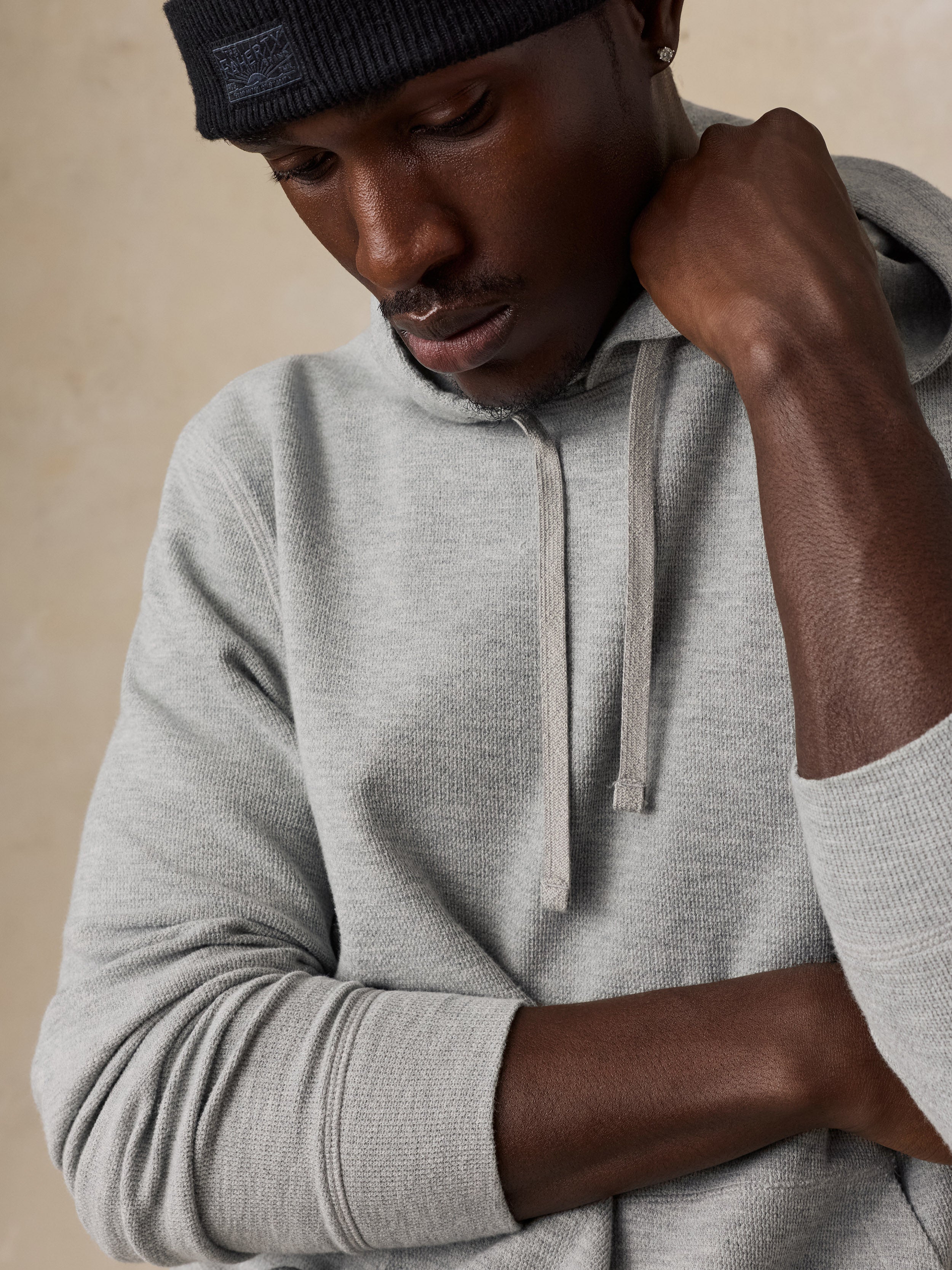 A close-up of a man wearing a light gray heathered hoodie with drawstrings and a dark ribbed beanie, posed against a neutral beige background.