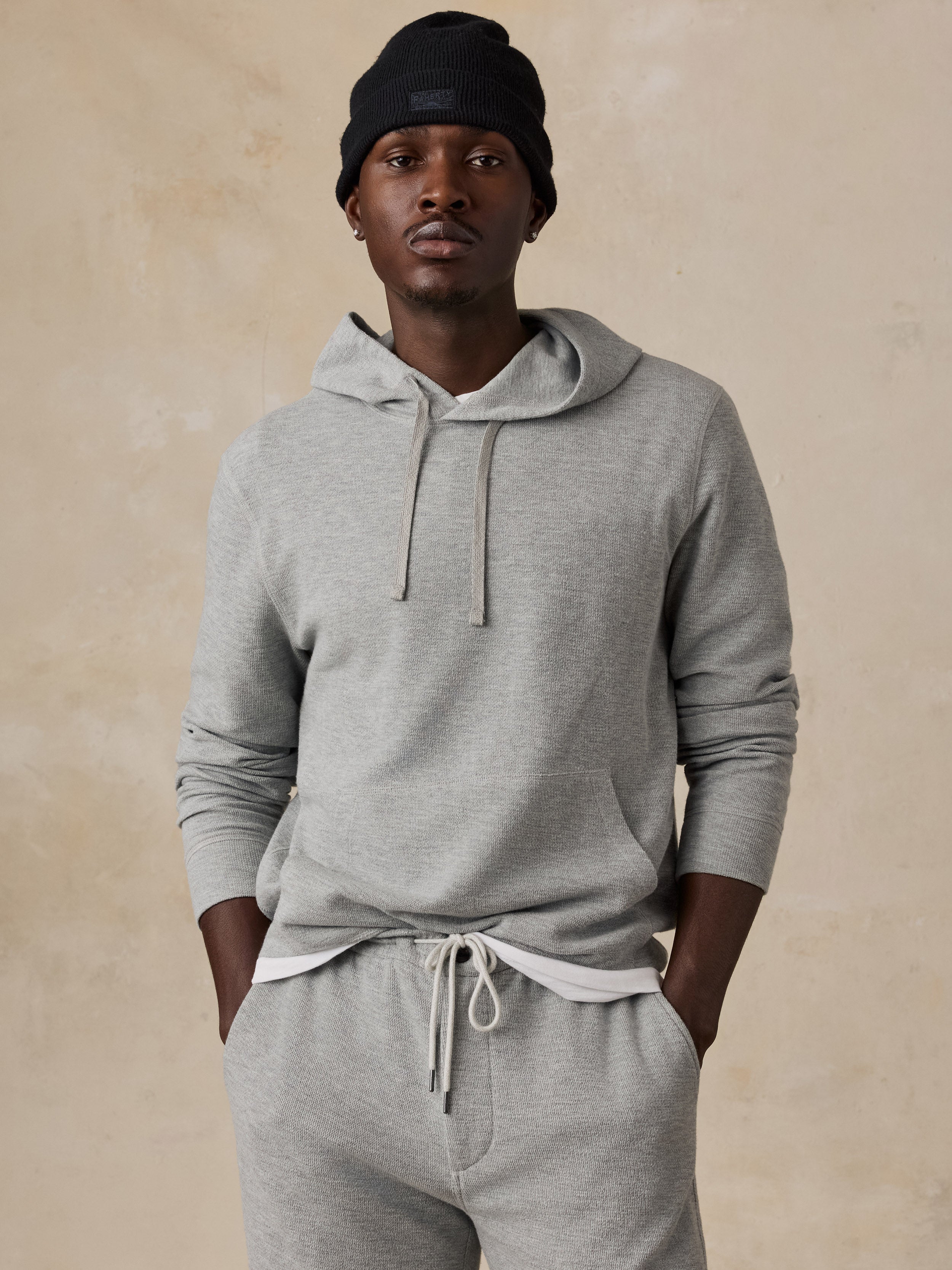 A man wearing a light gray heathered hoodie and matching jogger sweatpants with a drawstring waist, paired with a black beanie, standing against a textured beige background.