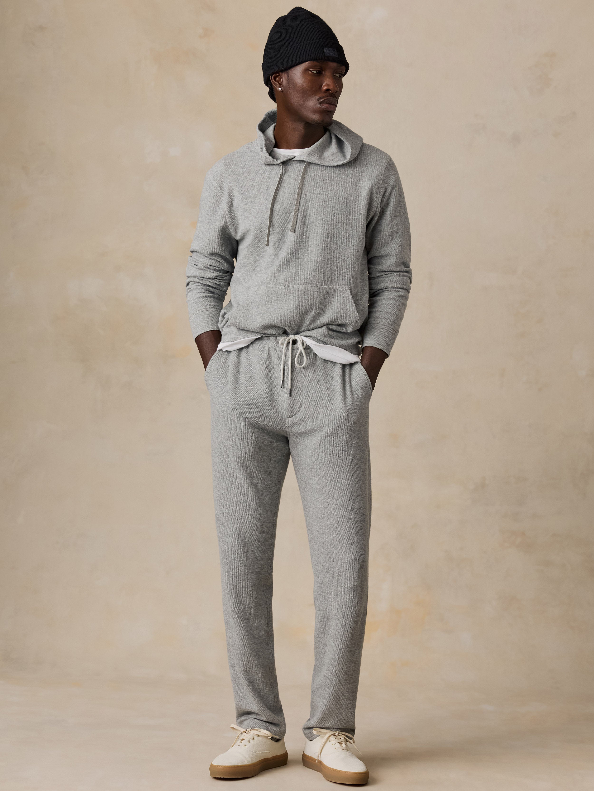A man modeling a light gray heathered two-piece loungewear set consisting of a drawstring hoodie and matching sweatpants, paired with a black beanie and off-white sneakers, against a textured beige studio backdrop.