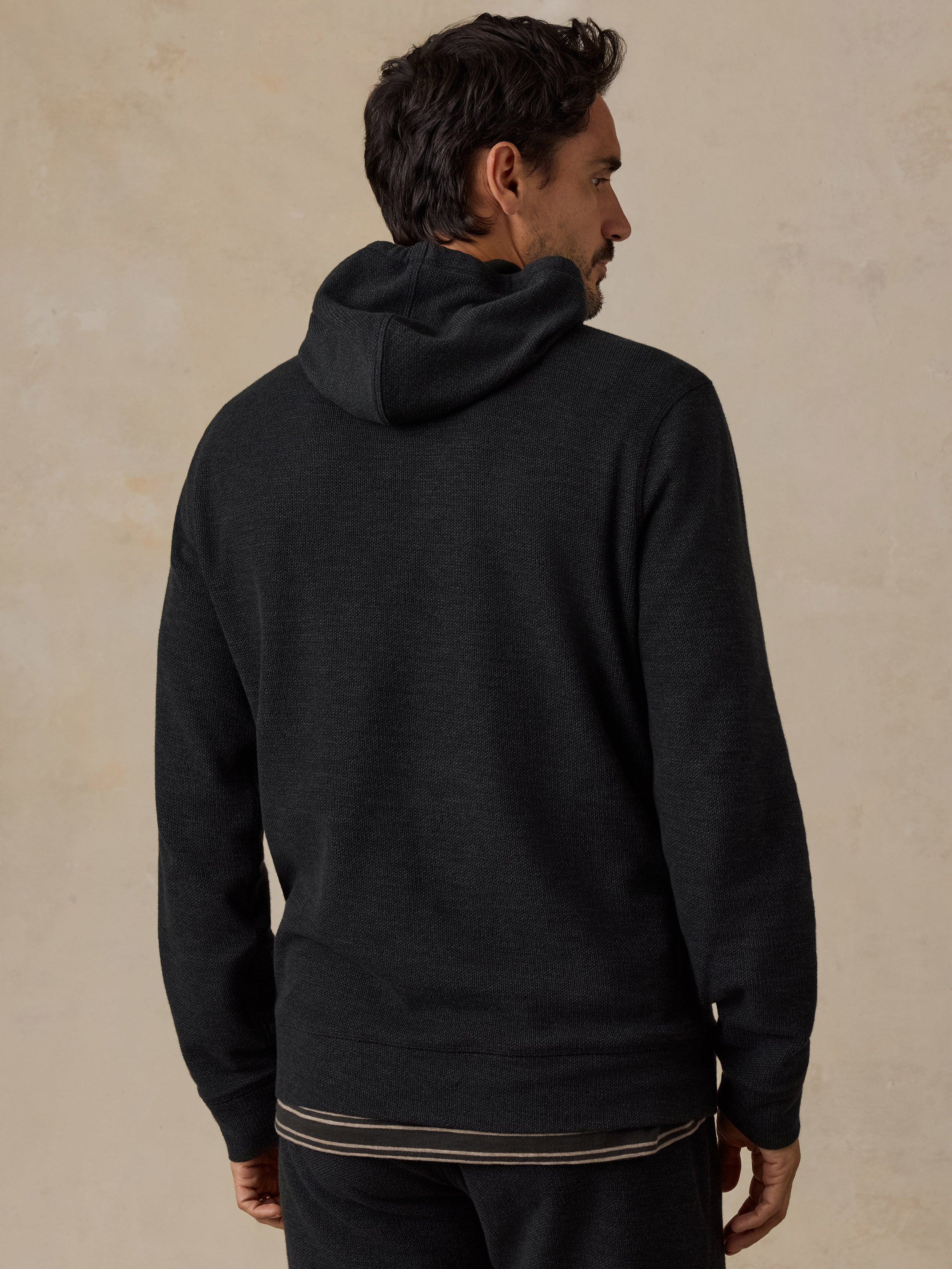 A man wearing a dark charcoal gray hooded sweatshirt, shown from the back and side profile against a textured beige background.