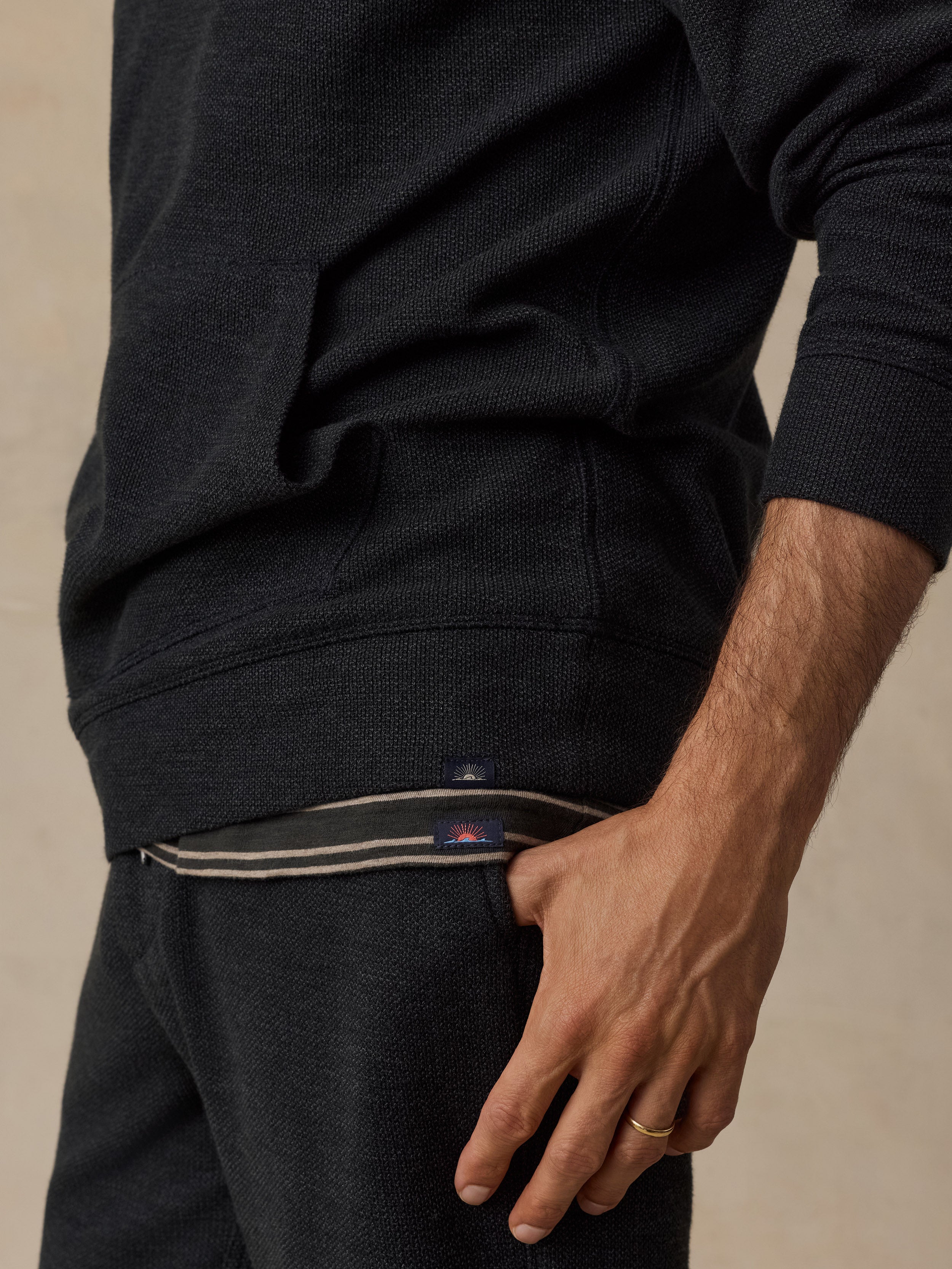 Close-up of a man wearing a dark charcoal knit hoodie and matching sweatpants, with the hand resting on the pocket area. A small Faherty label is visible on the hem.