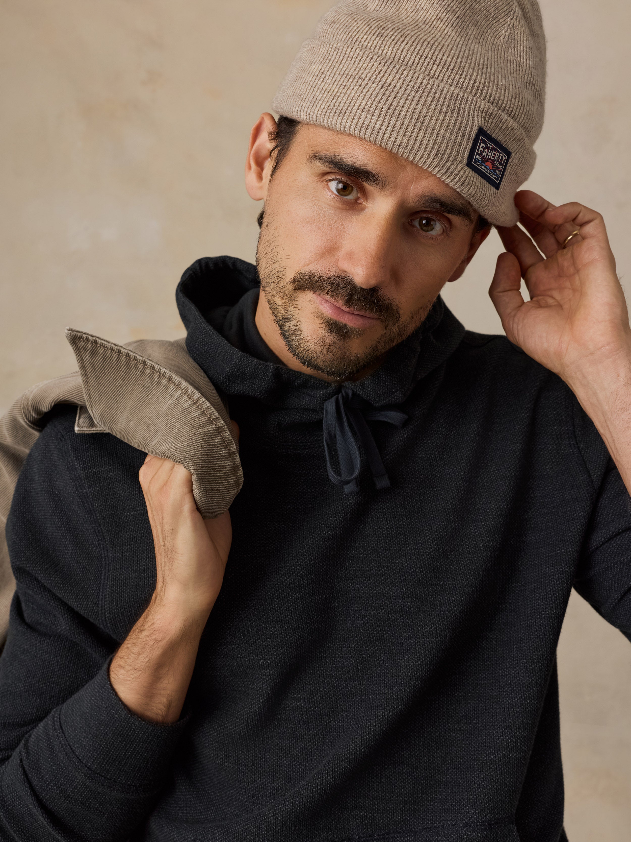 A man wearing a beige knit beanie with a small square Faherty brand patch on the cuff, a dark charcoal hoodie, and holding a matching beige knit garment over his shoulder, set against a textured beige background.