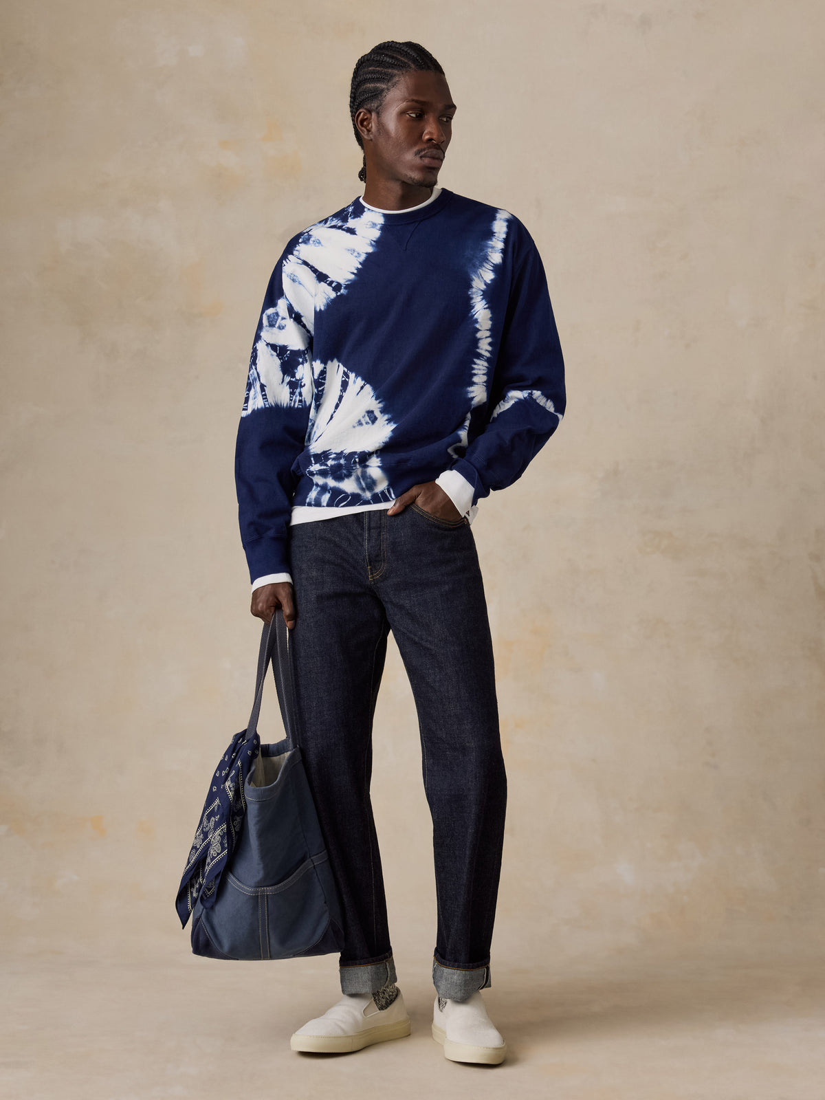 A man wearing a shibori dyed sweatshirt, blue jeans, and white sneakers holding a blue tote bag, image 2 of 6