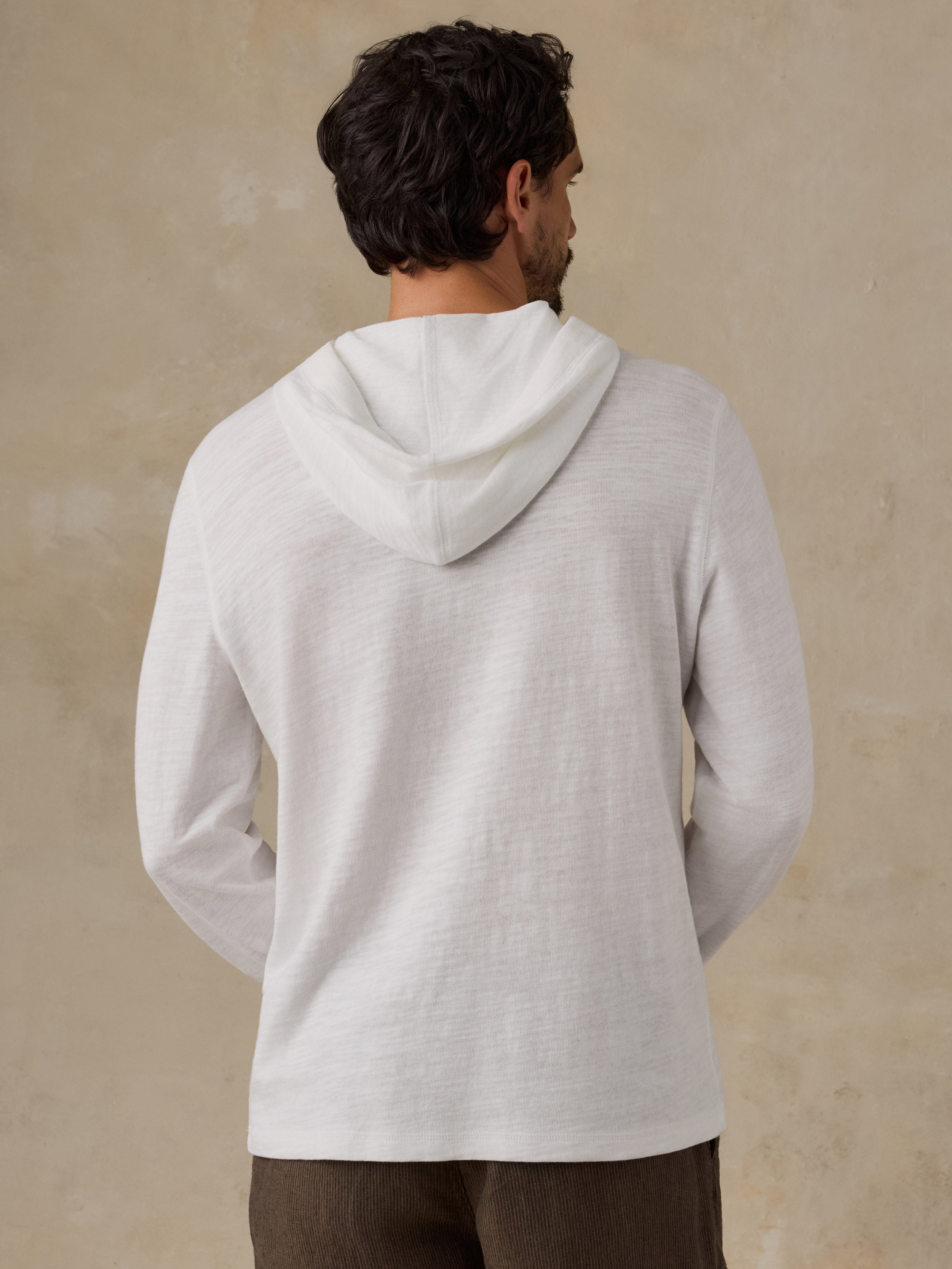 A man viewed from behind wearing a light off-white hooded sweater against a textured beige wall.