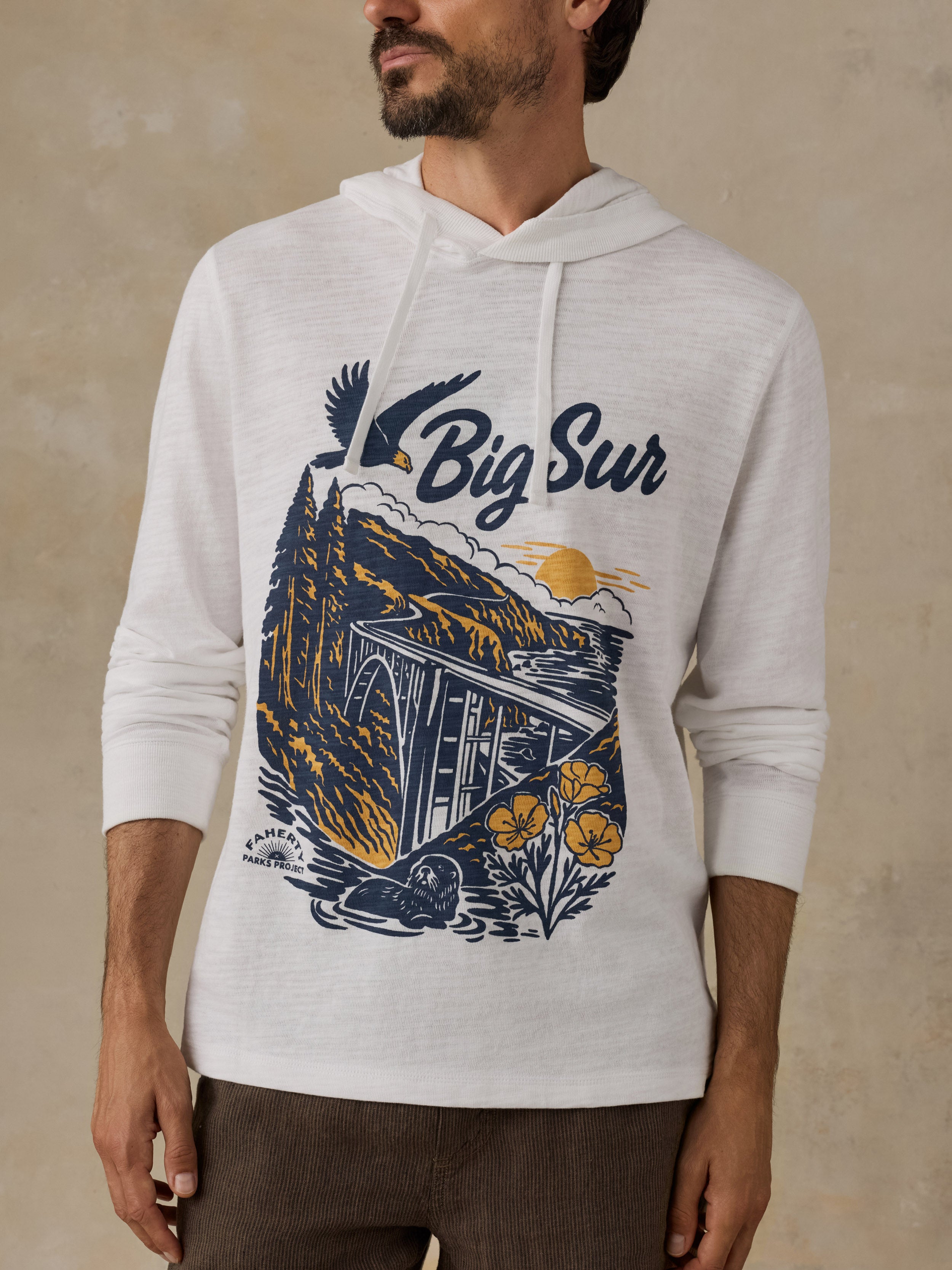 A man wearing a white heathered pullover hoodie with a large 'Big Sur' graphic print in navy blue and yellow on the front, paired with brown pants, set against a textured beige background.