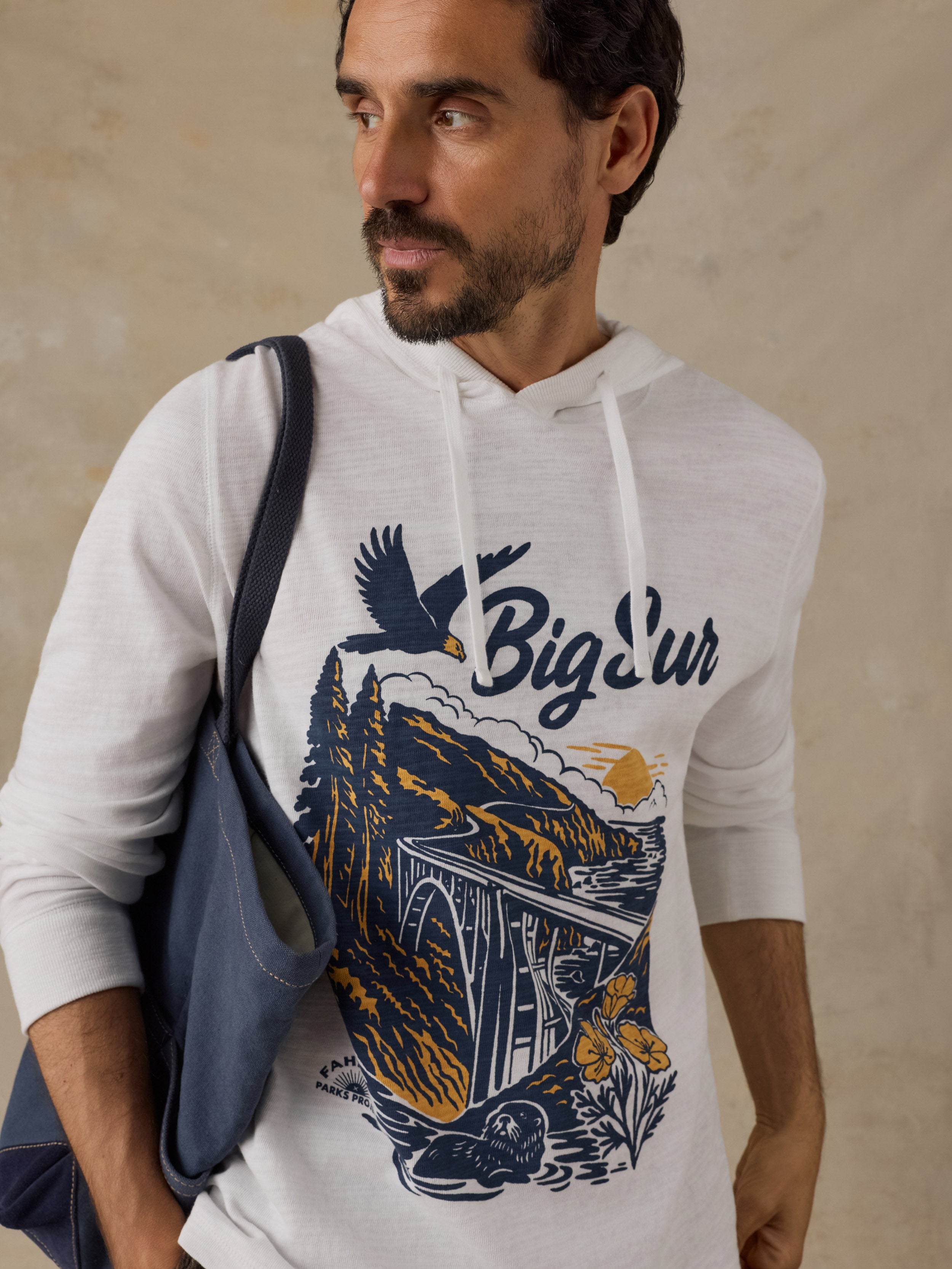A man wearing a white heathered hoodie with a large navy and orange graphic print on the front that reads 'Big Sur' and shows a coastal landscape scene. He is carrying a blue shoulder bag against a textured beige wall.