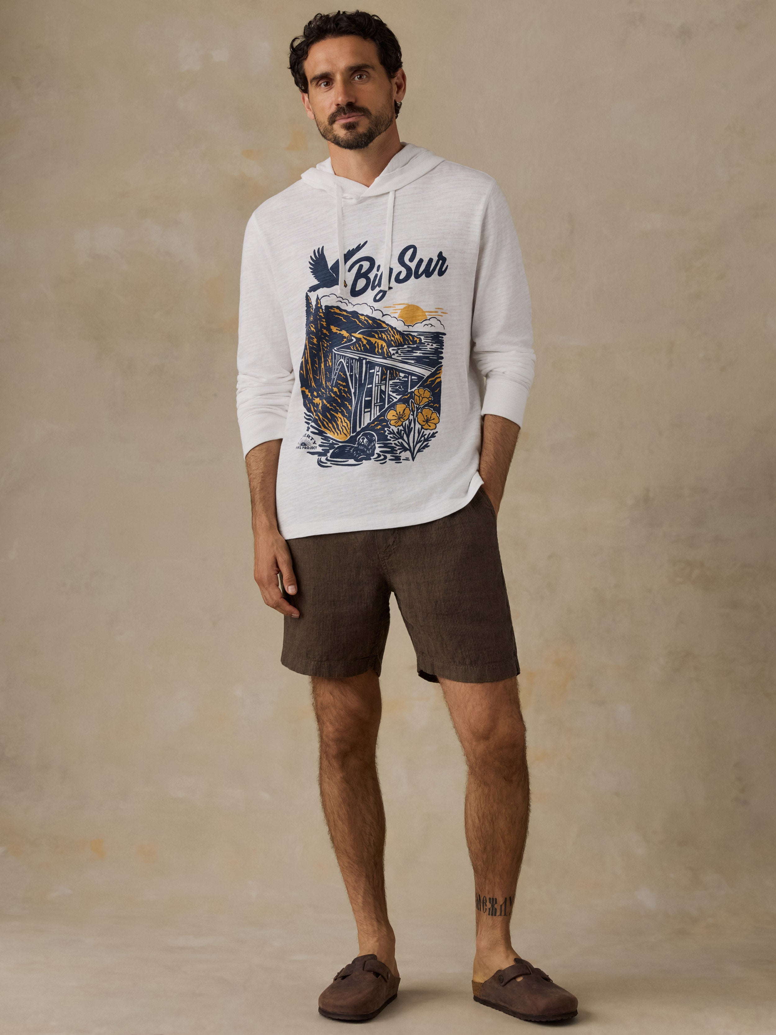 A man wearing a white long-sleeve hoodie with a blue and yellow graphic print that reads 'Big Sur', paired with brown shorts and brown slip-on shoes, standing against a textured beige studio backdrop.