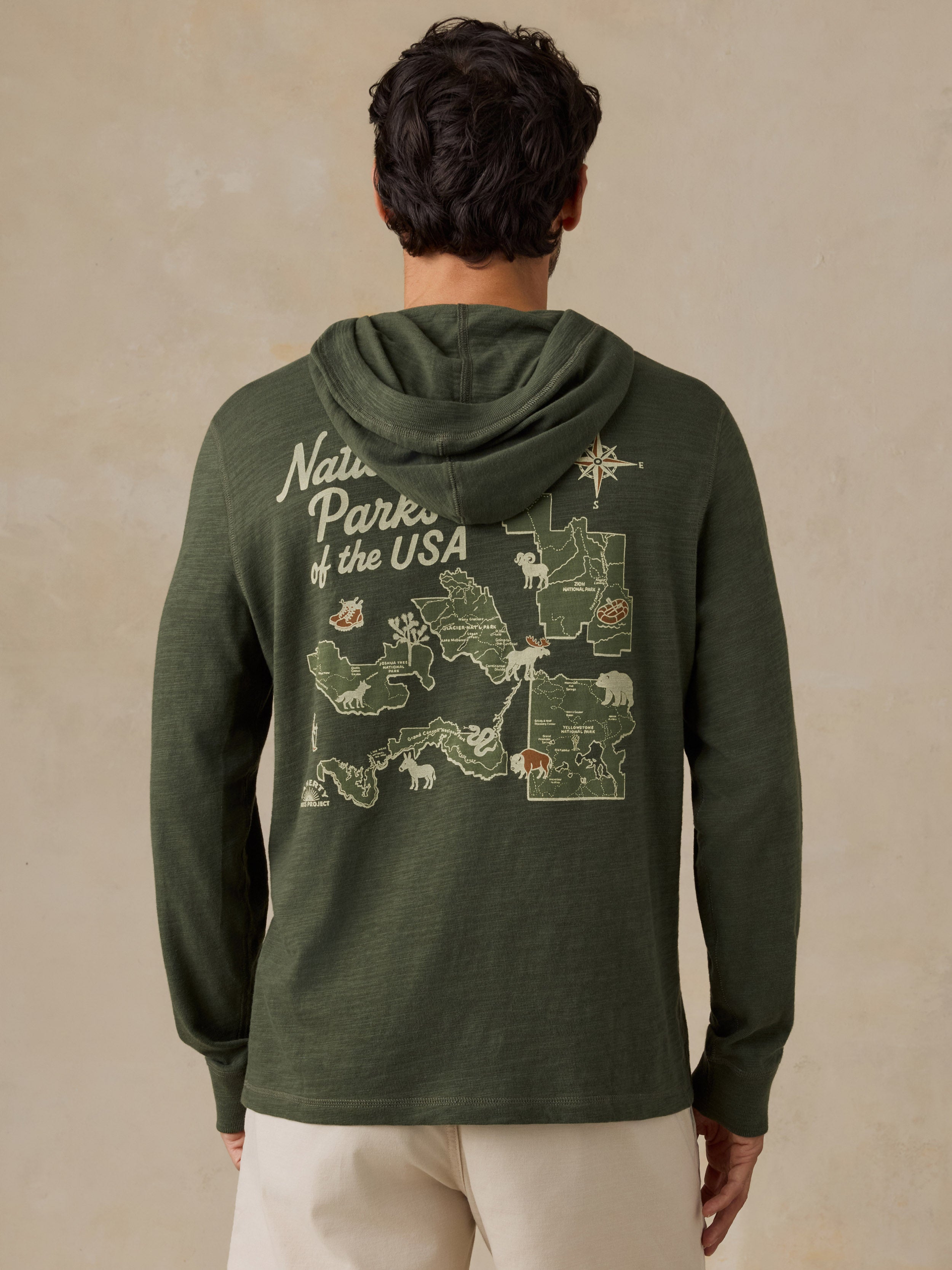 A man viewed from behind wearing an olive green hooded sweatshirt with a large 'National Parks of the USA' map graphic printed on the back, paired with light beige shorts, against a textured beige background.