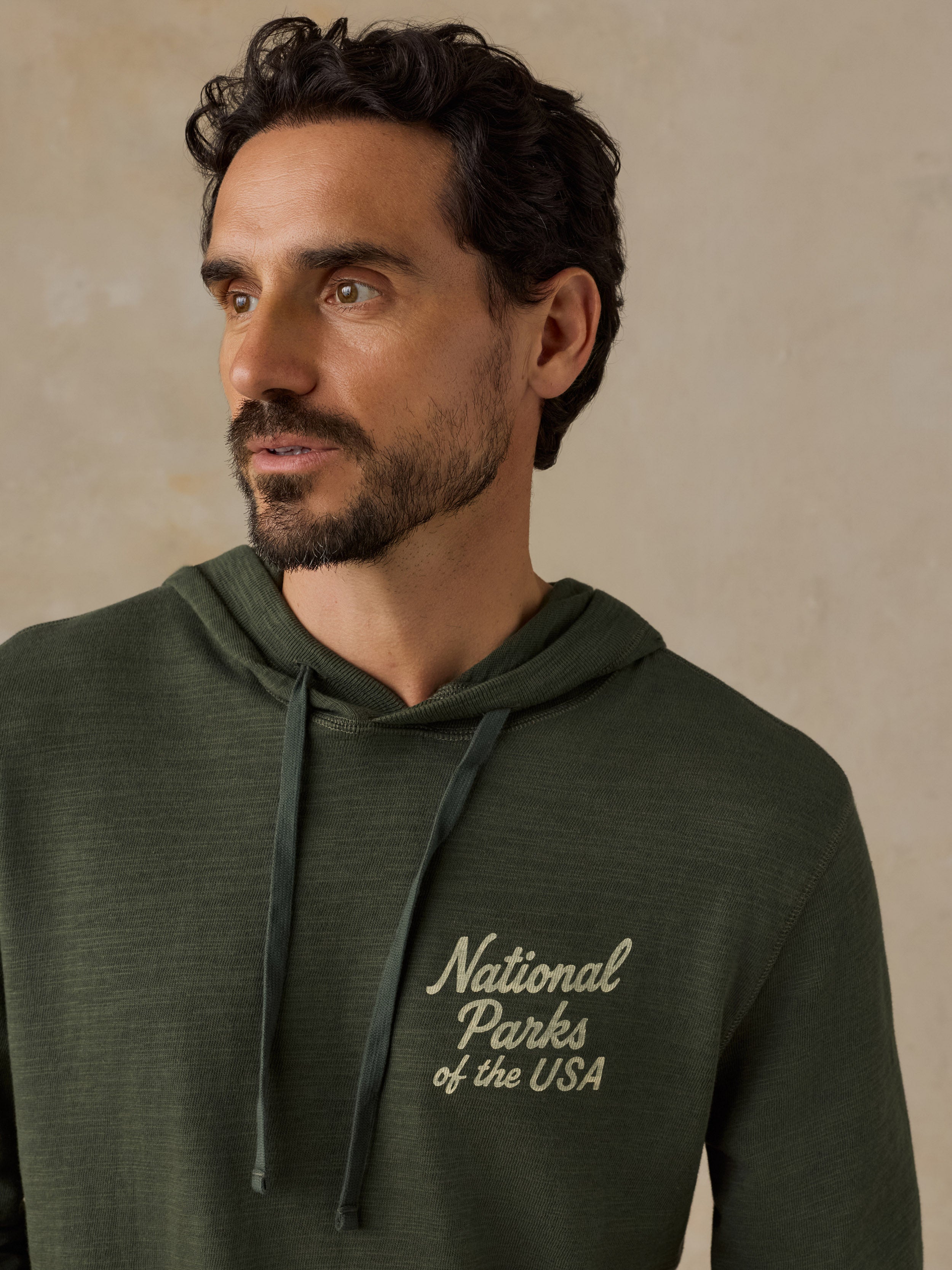 A man wearing an olive green hooded sweatshirt with 'National Parks of the USA' printed in cream on the left chest, set against a textured beige background.