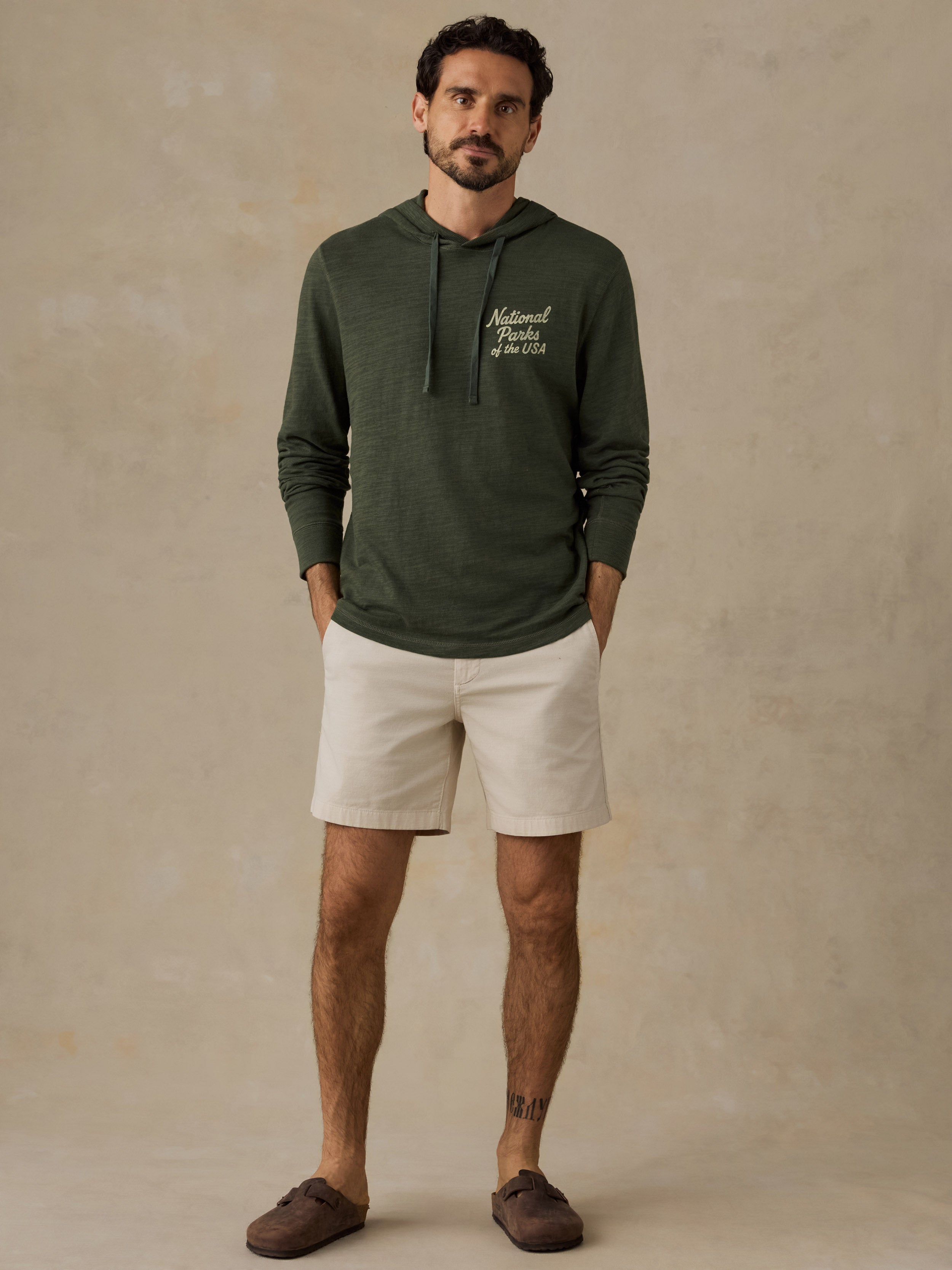 A man wearing an olive green hooded sweatshirt with white embroidered text on the left chest, paired with off-white shorts and brown slip-on shoes, standing against a textured beige studio backdrop.