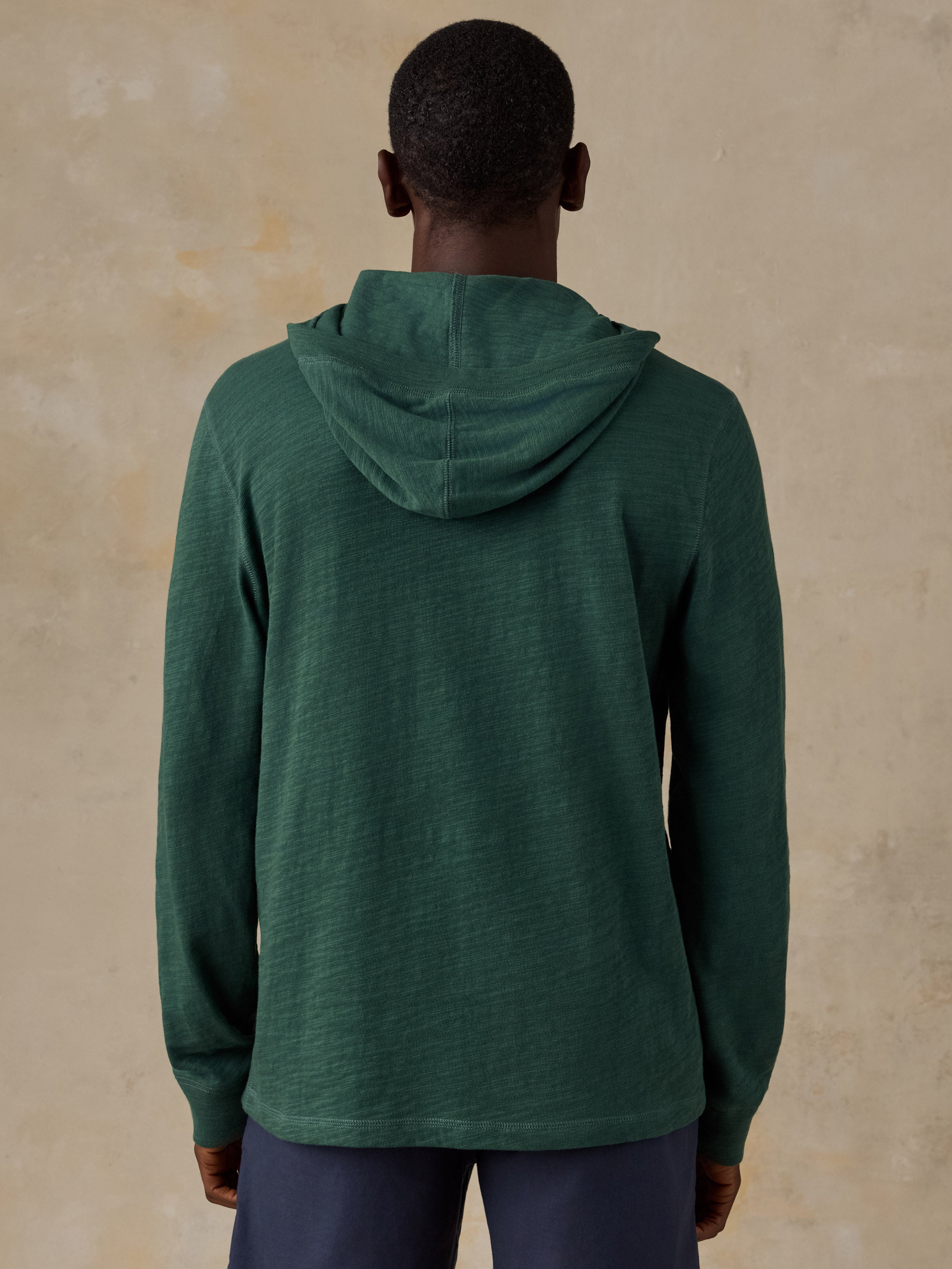A man viewed from behind wearing a dark green hooded long-sleeve shirt and navy pants, standing against a textured beige wall.