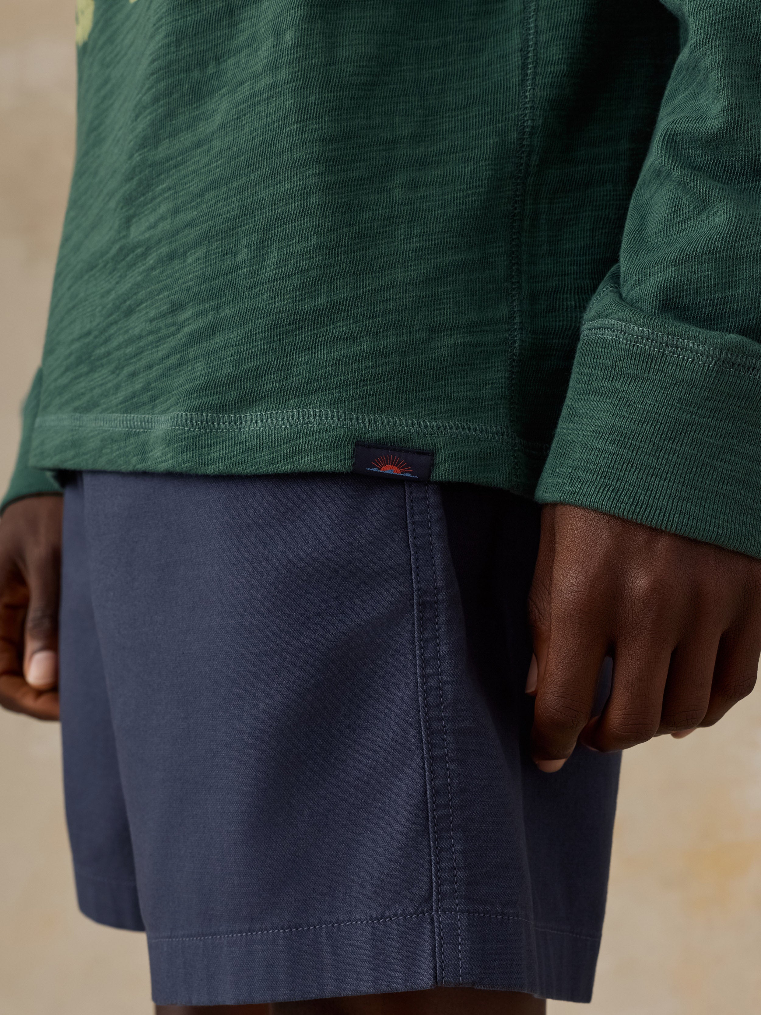 Close-up of a man wearing a dark green long-sleeve hoodie and navy blue shorts, with a small Faherty brand sun and waves tag visible on the hem of the hoodie.
