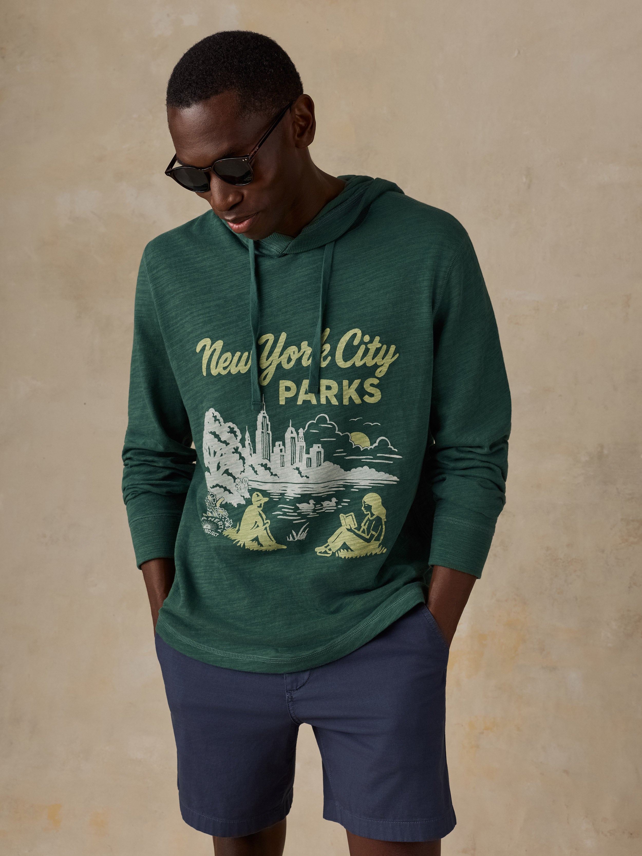 A man wearing a dark green heathered hoodie with a vintage-style 'New York City Parks' graphic on the front, paired with navy blue shorts, set against a textured beige background.