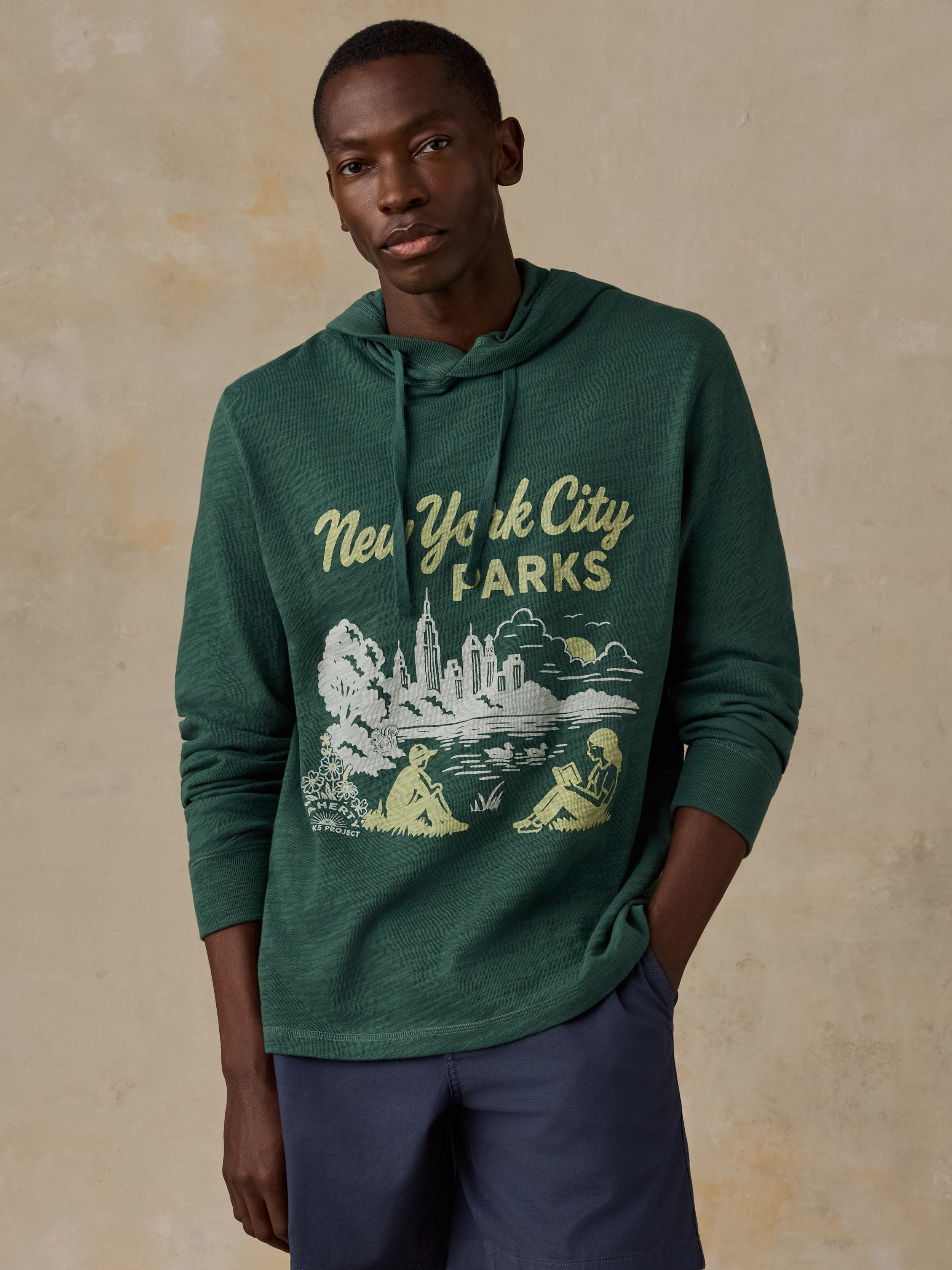 A man wearing a dark green heathered pullover hoodie with a 'New York City Parks' graphic on the front, paired with navy blue pants, standing against a textured beige background.