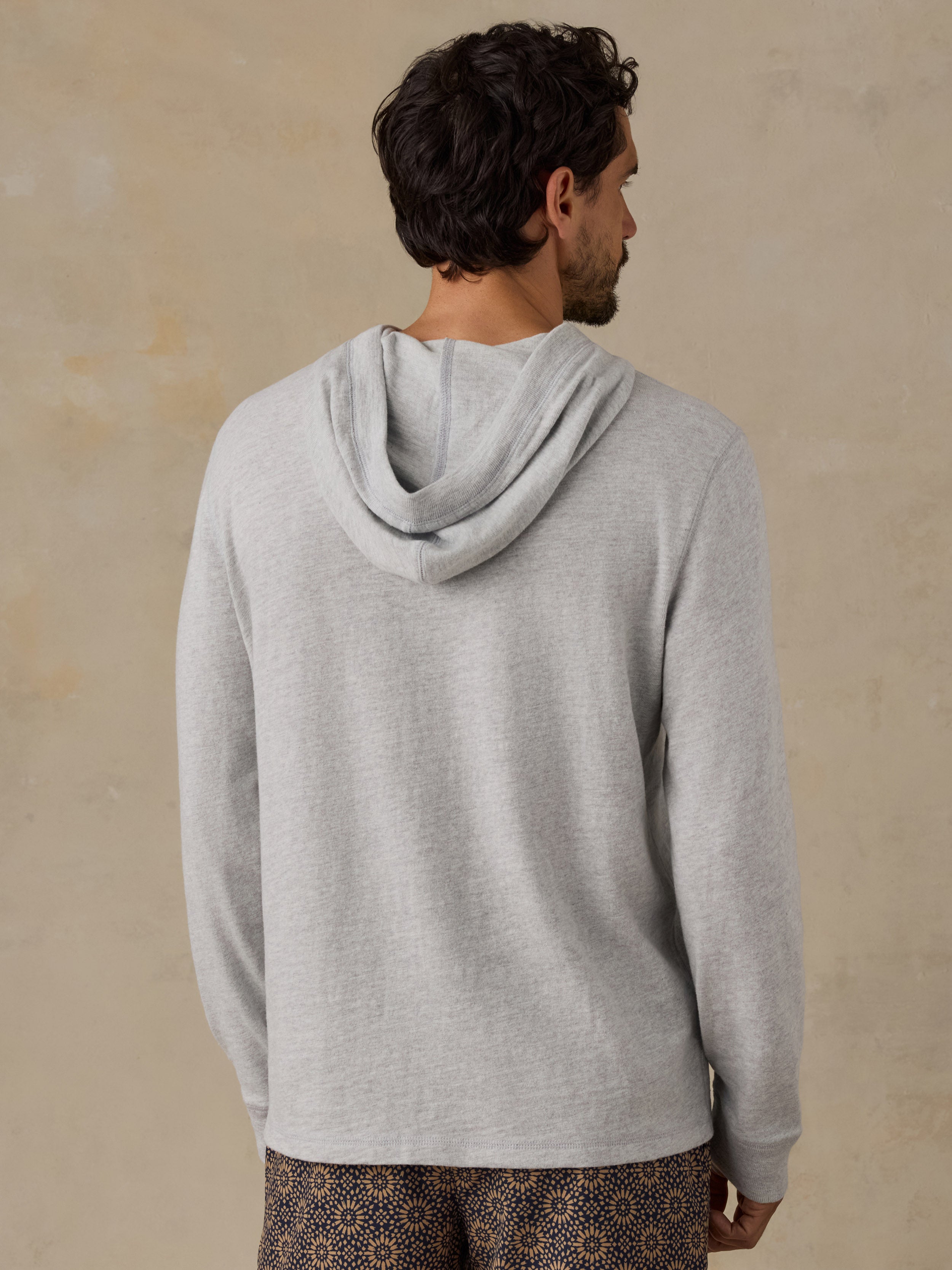 A man viewed from behind wearing a light gray heathered hooded sweatshirt and patterned brown shorts, standing against a textured beige wall.