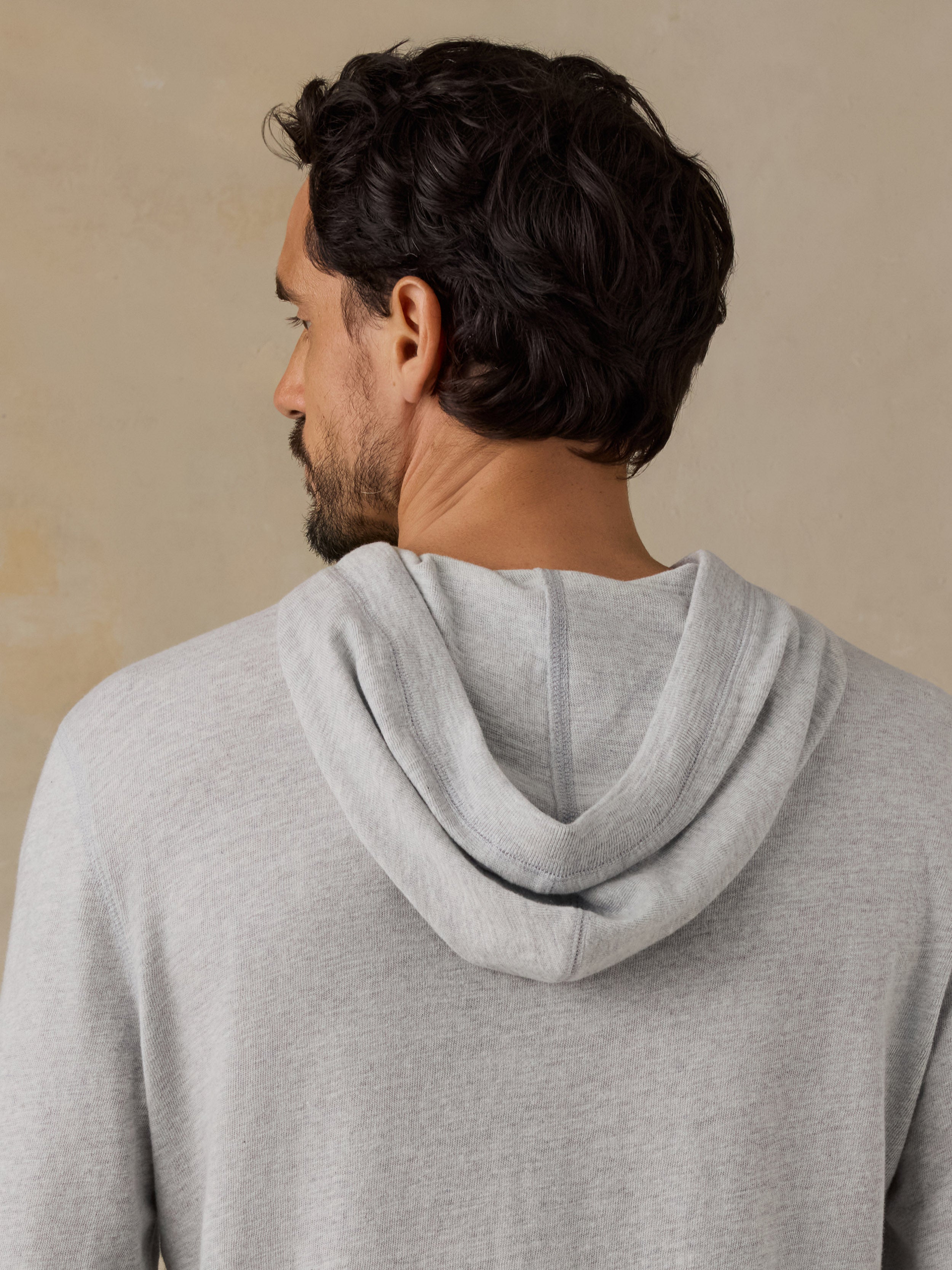 A man shown from the back wearing a light gray heathered hooded sweatshirt against a textured beige background.