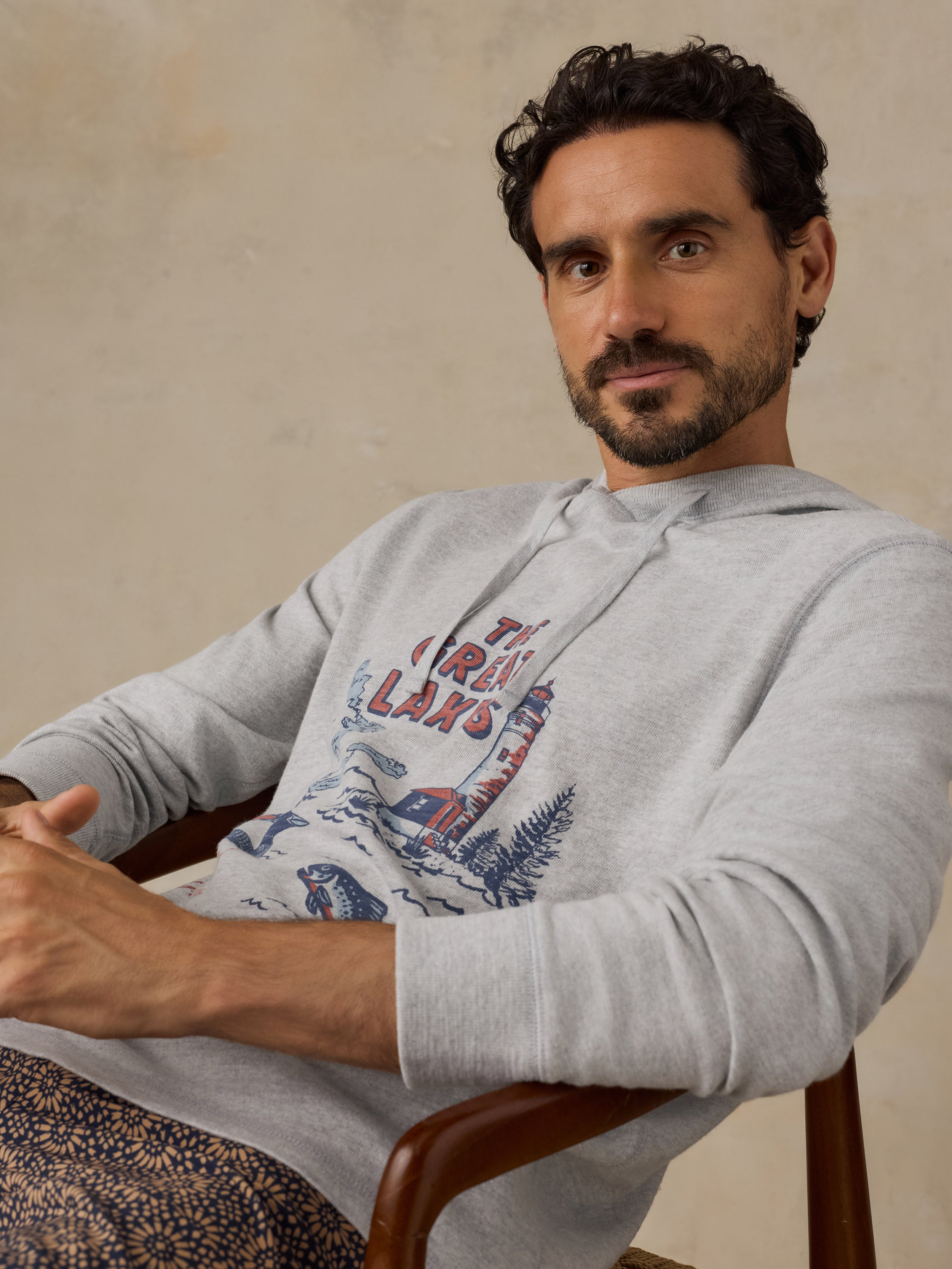 A man wearing a light gray hoodie with a red and blue graphic print that reads 'THE GREAT LAKES' sits in a wooden chair against a textured beige wall.