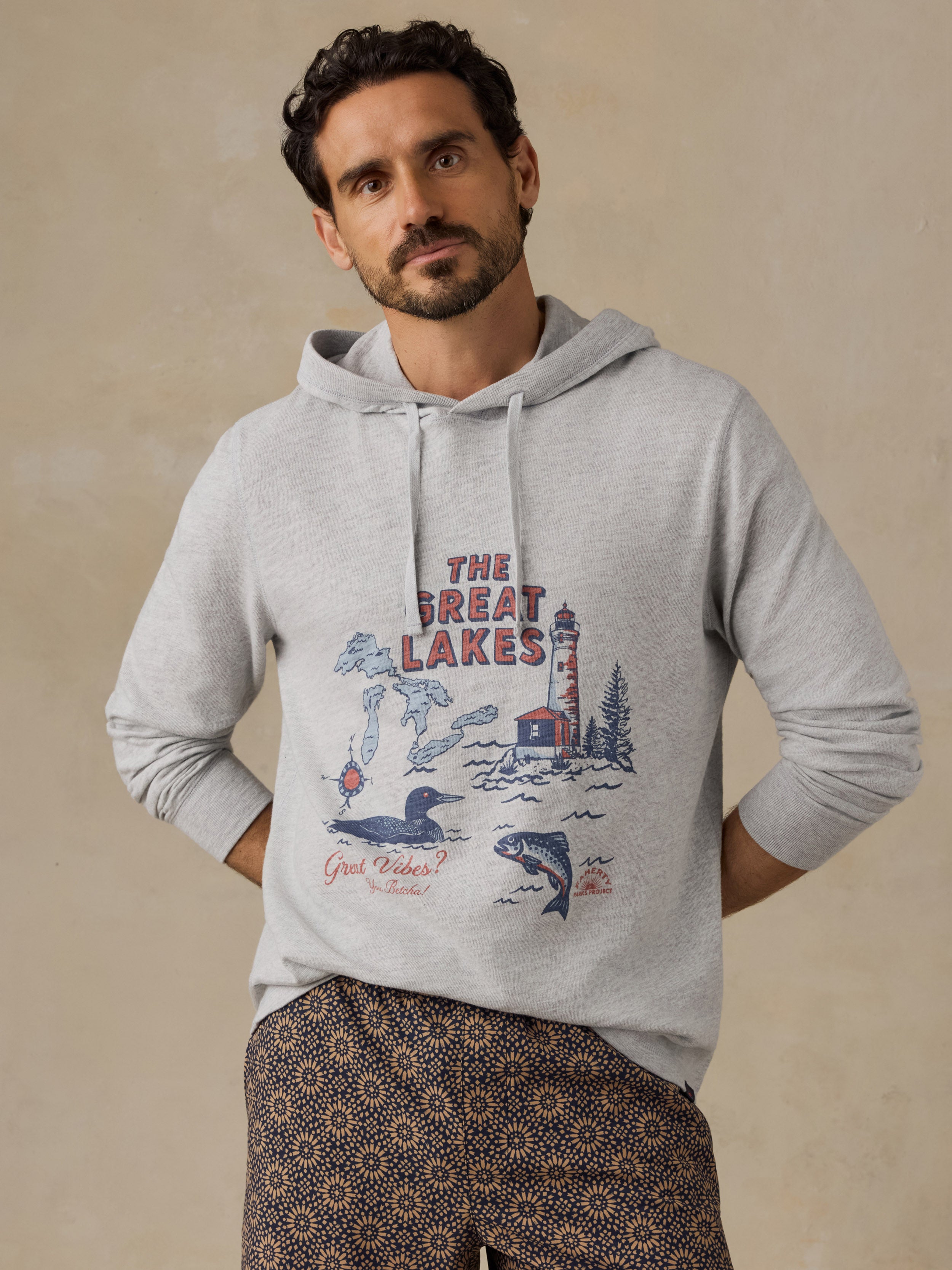 A man wearing a light gray heathered hoodie with a vintage-style 'The Great Lakes' graphic print on the front, paired with patterned shorts, standing against a textured beige background.