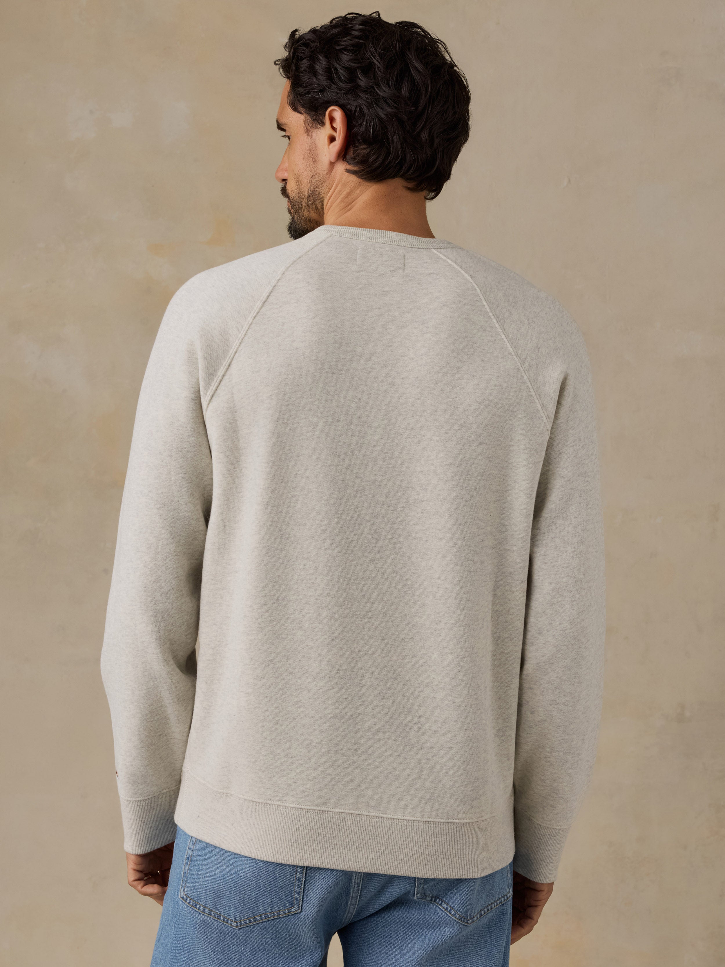 A man viewed from behind wearing a light beige crewneck sweatshirt and blue jeans, standing against a textured beige wall.