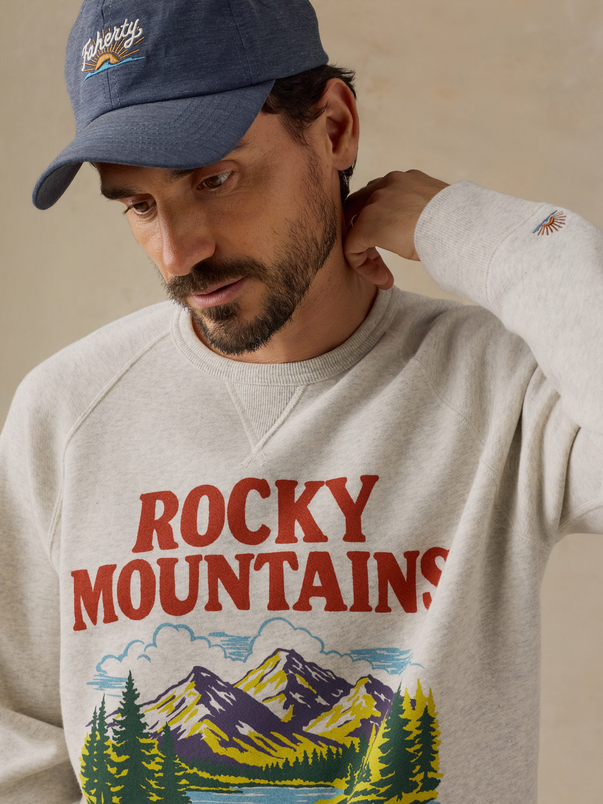 A man wearing a light gray crewneck sweatshirt with a 'ROCKY MOUNTAINS' graphic print in red, green, yellow, and blue, along with a navy blue embroidered cap.
