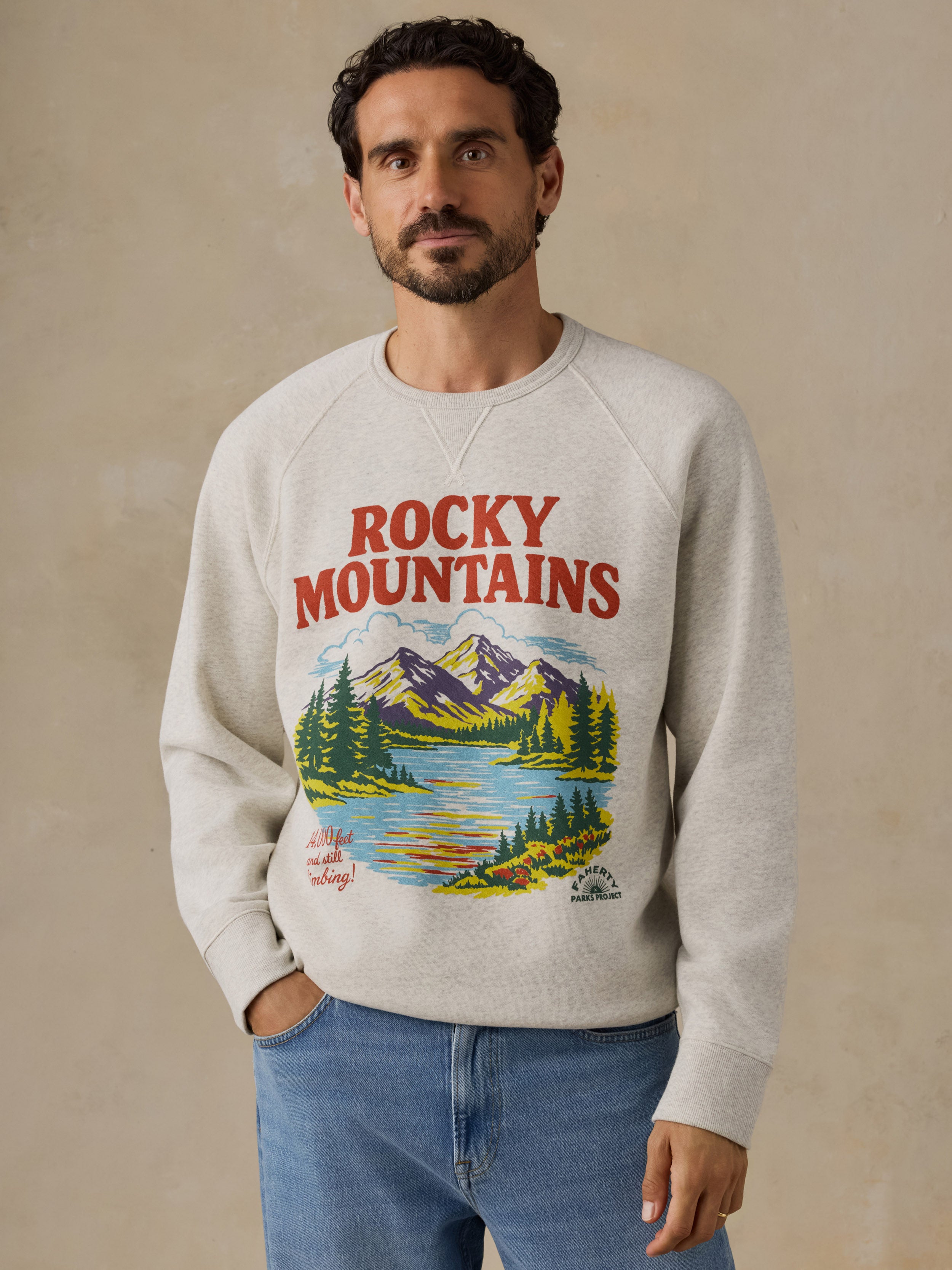 A man wearing a light beige crewneck sweatshirt with a colorful 'Rocky Mountains' graphic print on the front, paired with blue jeans, set against a neutral beige background.