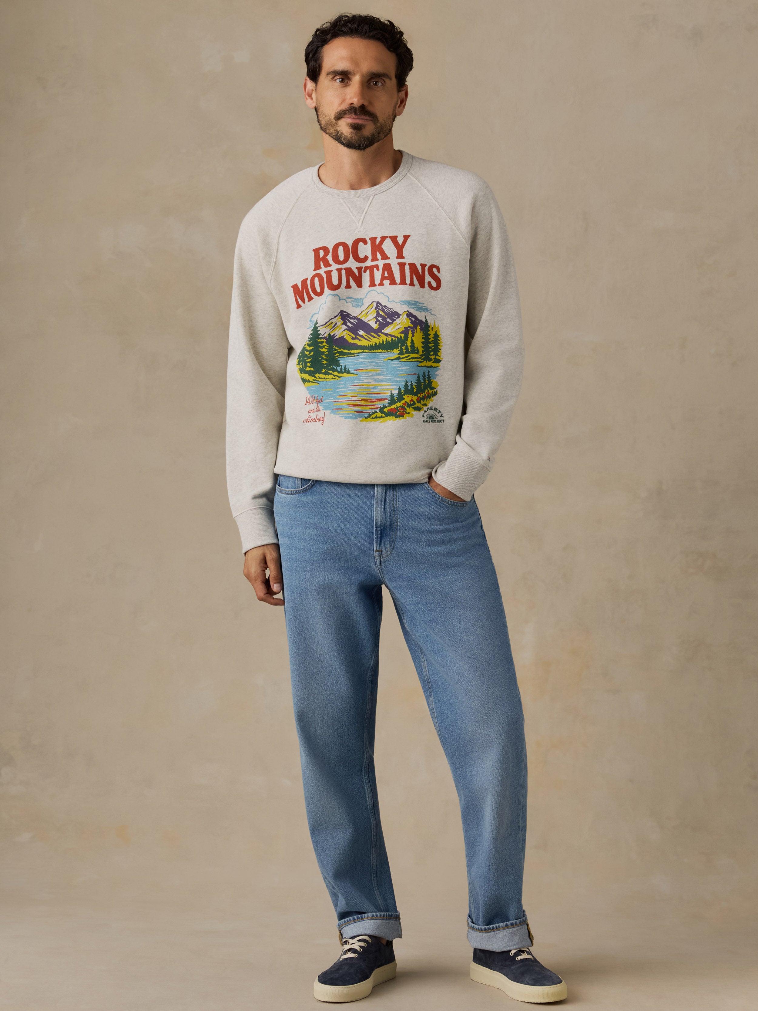 A man wearing a light beige crewneck sweatshirt with a colorful 'Rocky Mountains' graphic print, paired with blue jeans and navy sneakers, standing against a textured beige studio backdrop.