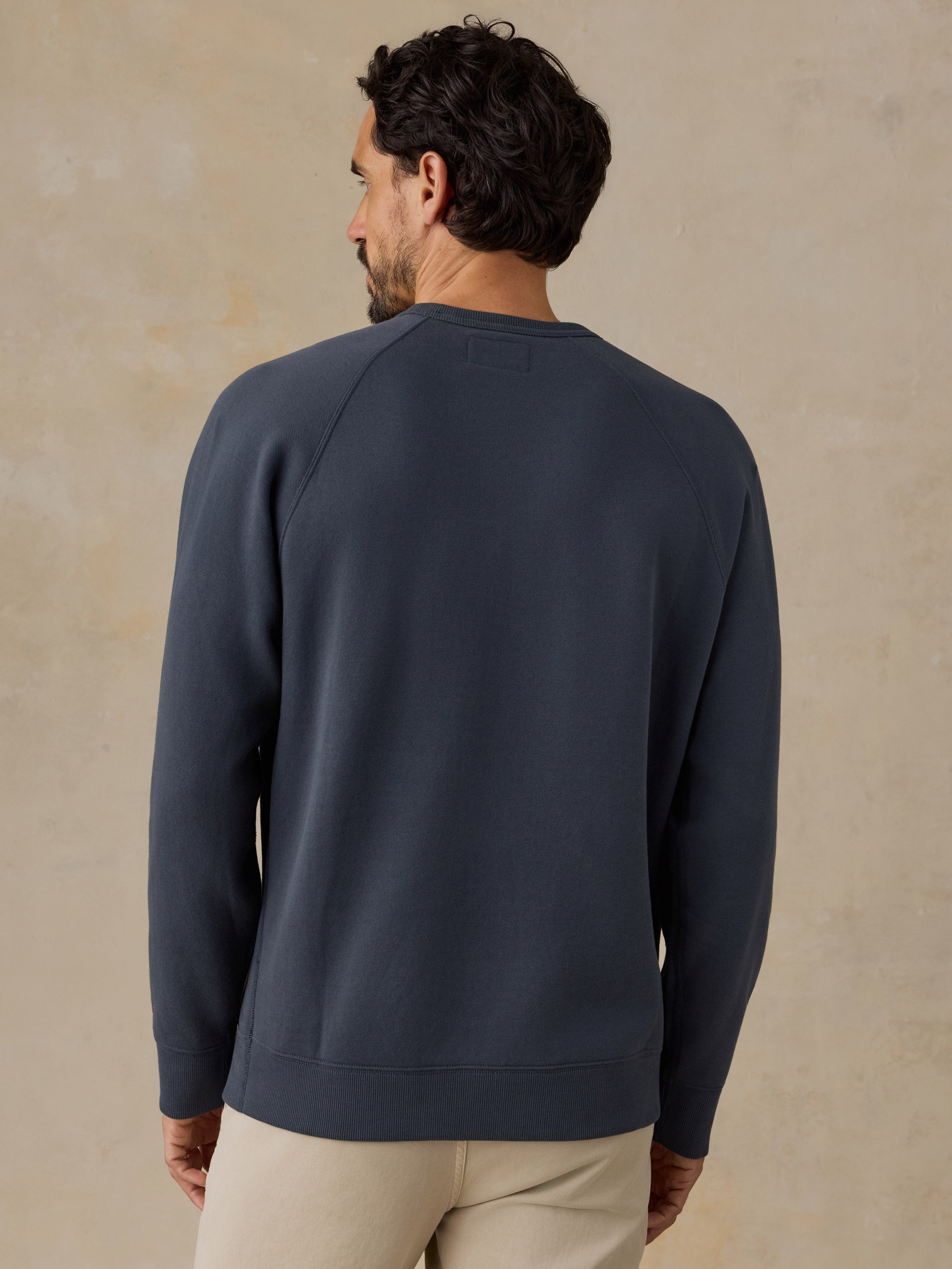 A man wearing a navy blue crewneck sweatshirt, shown from the back against a textured beige background.