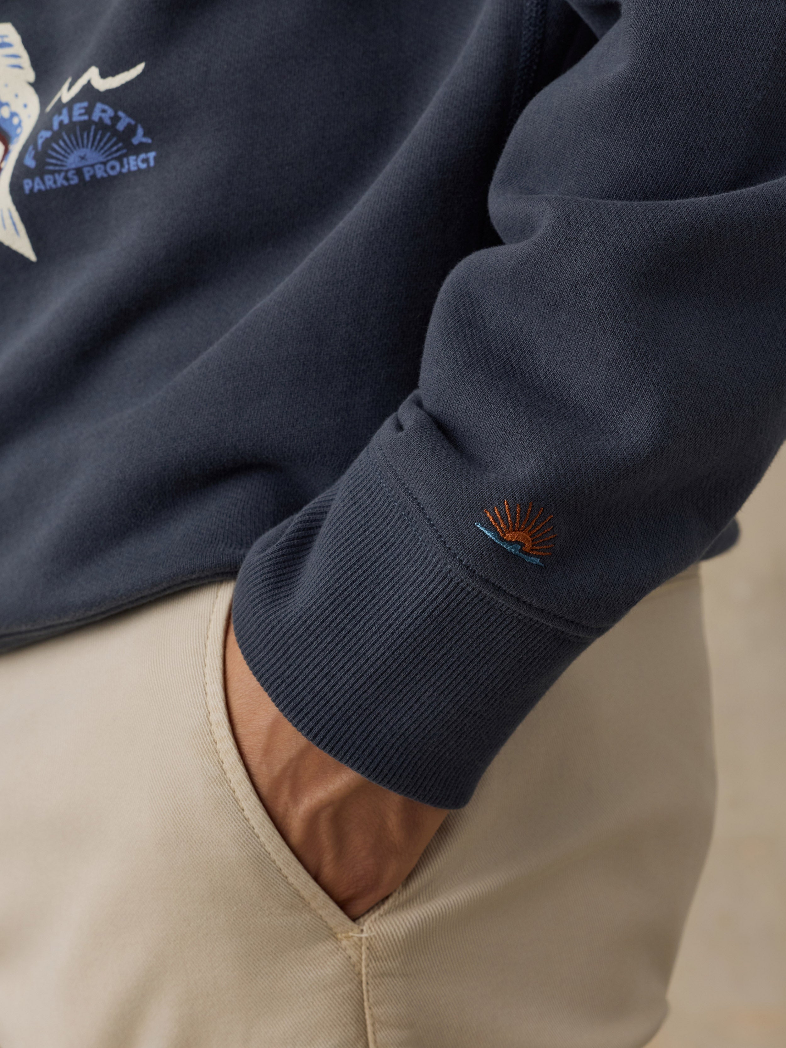 Close-up of a navy blue sweatshirt worn with beige pants, showing the ribbed cuff with a small orange and blue embroidered Faherty brand logo near the sleeve cuff