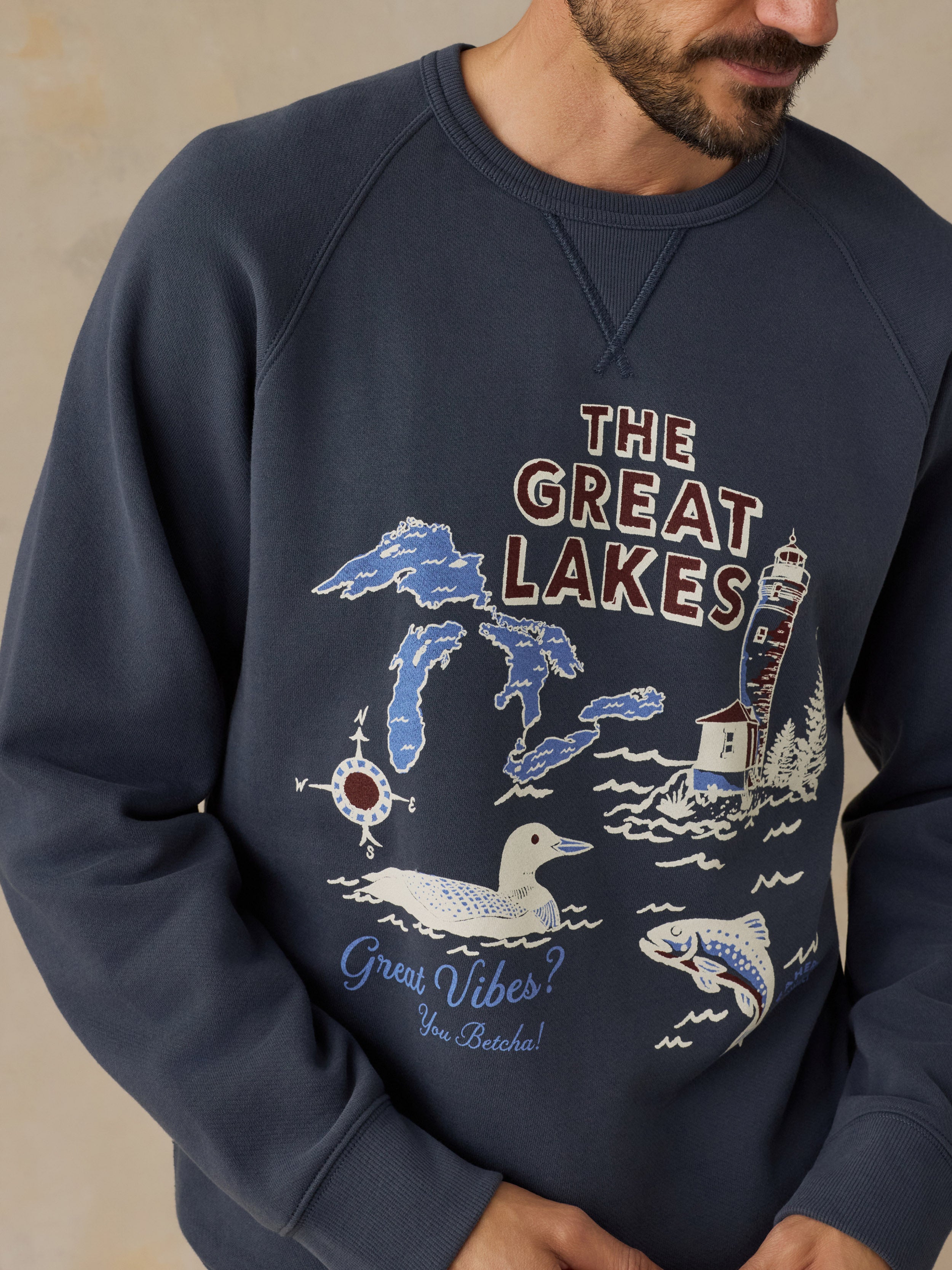 A man wearing a navy blue crewneck sweatshirt with a graphic print of the Great Lakes, including Michigan, a lighthouse, a duck, a fish, and the text 'The Great Lakes' and 'Great Vibes You Deserve!'.