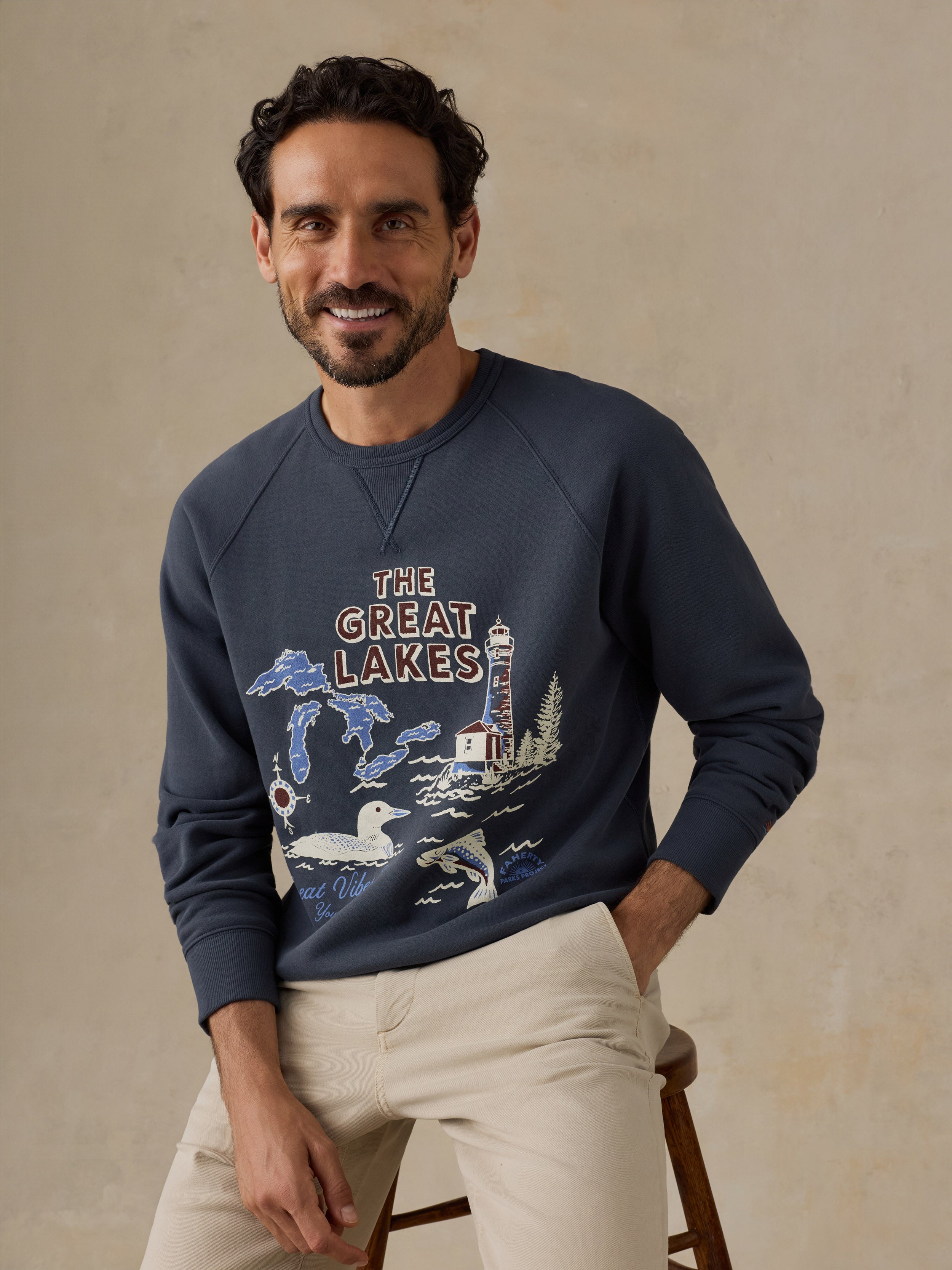 A man wearing a navy blue crewneck sweatshirt with a 'The Great Lakes' graphic print, paired with beige trousers, seated on a wooden stool against a textured beige wall.