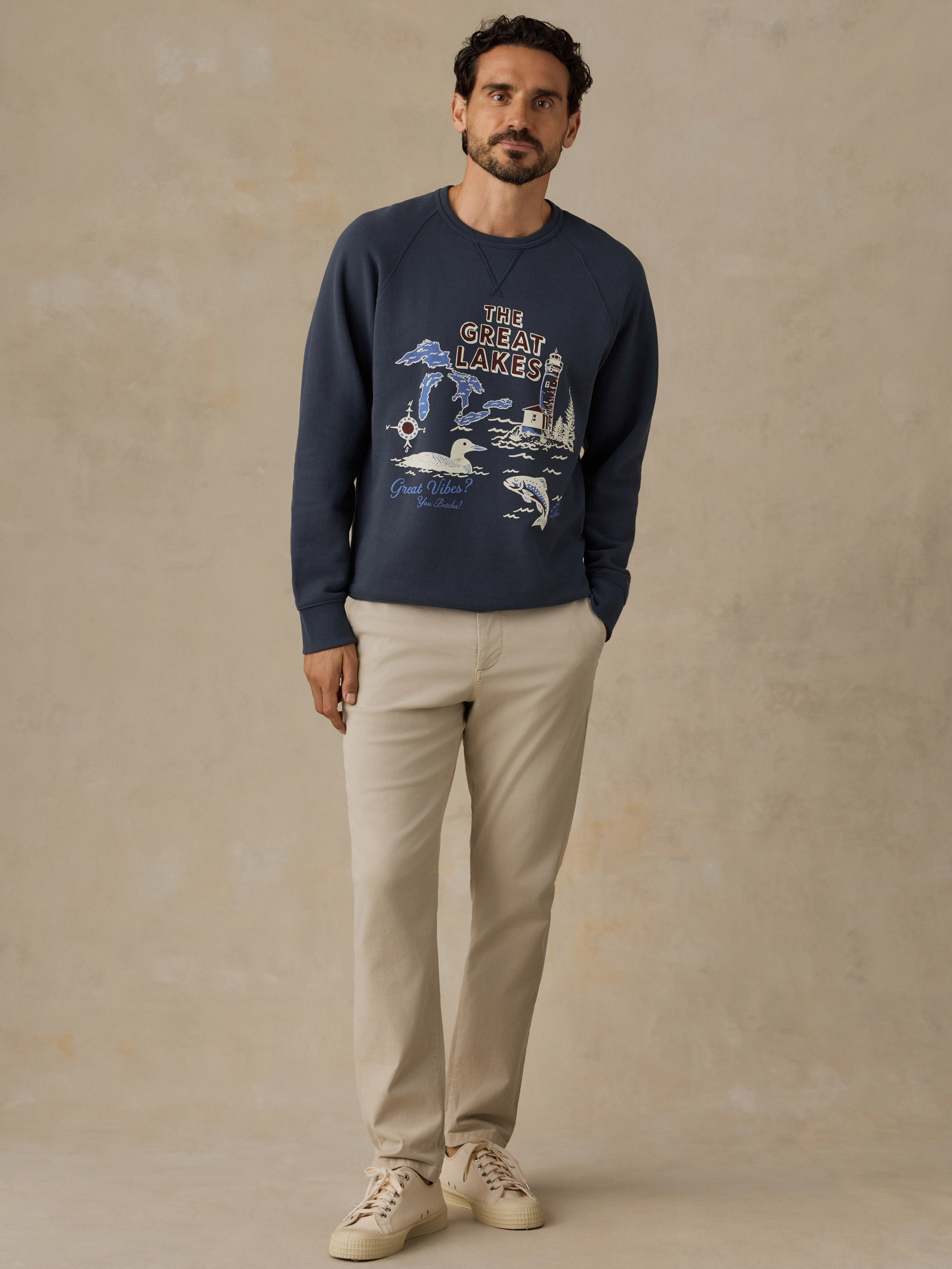A man wearing a navy blue crewneck sweatshirt with a 'The Great Lakes' graphic, paired with beige trousers and beige sneakers, standing against a textured beige studio backdrop.
