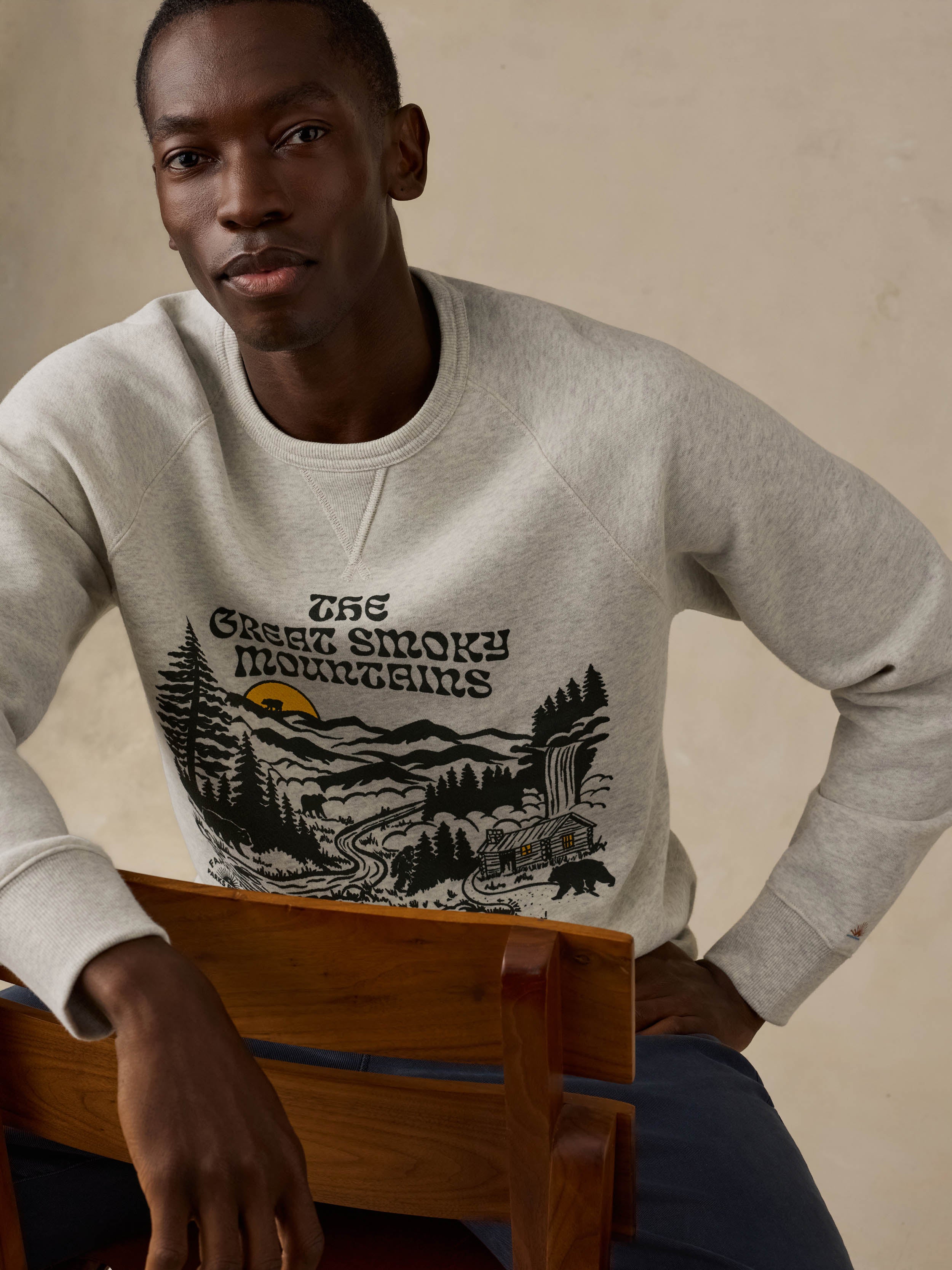 Man sitting on a wooden chair wearing a gray sweatshirt with a nature graphic and text on a beige background
