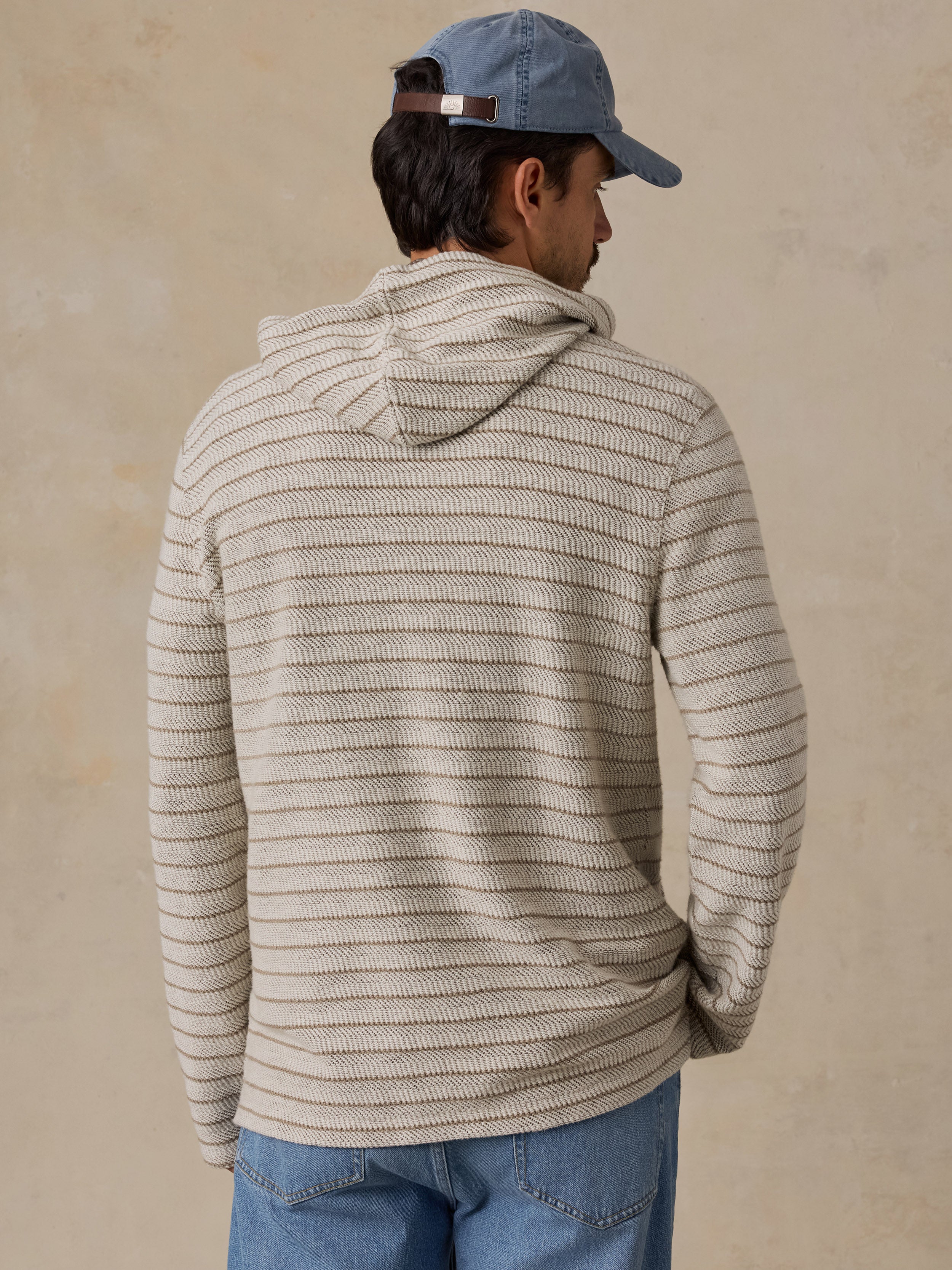A man wearing a beige and gray horizontally striped knit hoodie, blue jeans, and a blue baseball cap, shown from a back-three-quarter view against a textured beige wall.