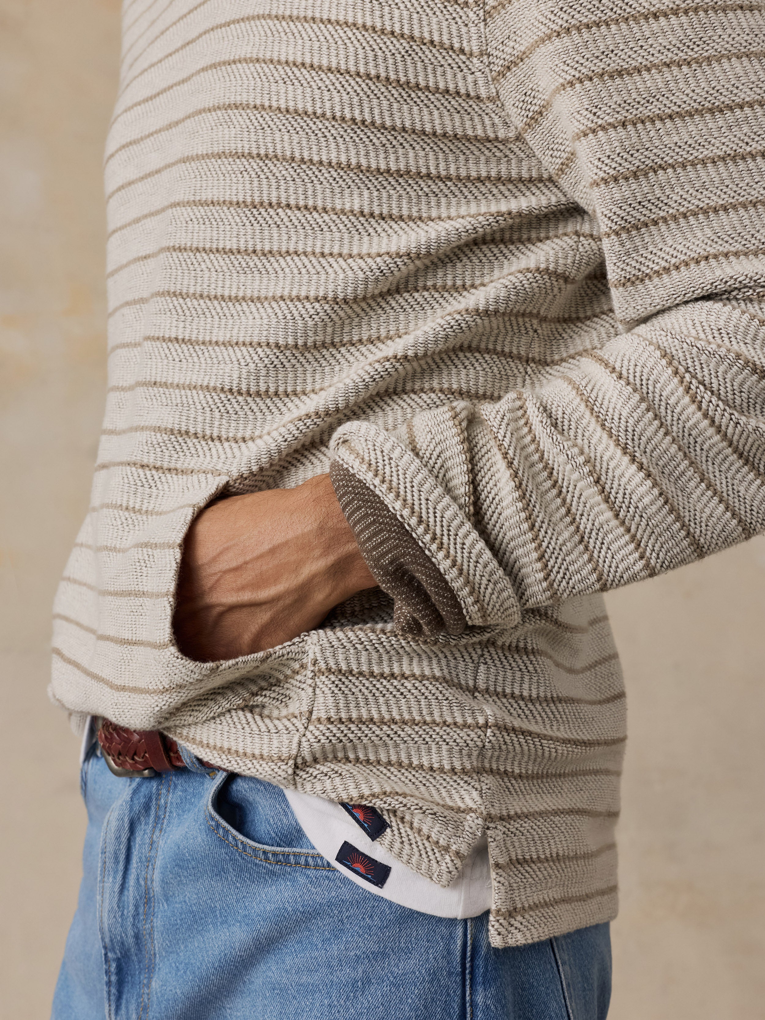 Close-up of a man wearing a beige and brown horizontally striped ribbed knit sweater with a pocket, tucked into blue jeans.