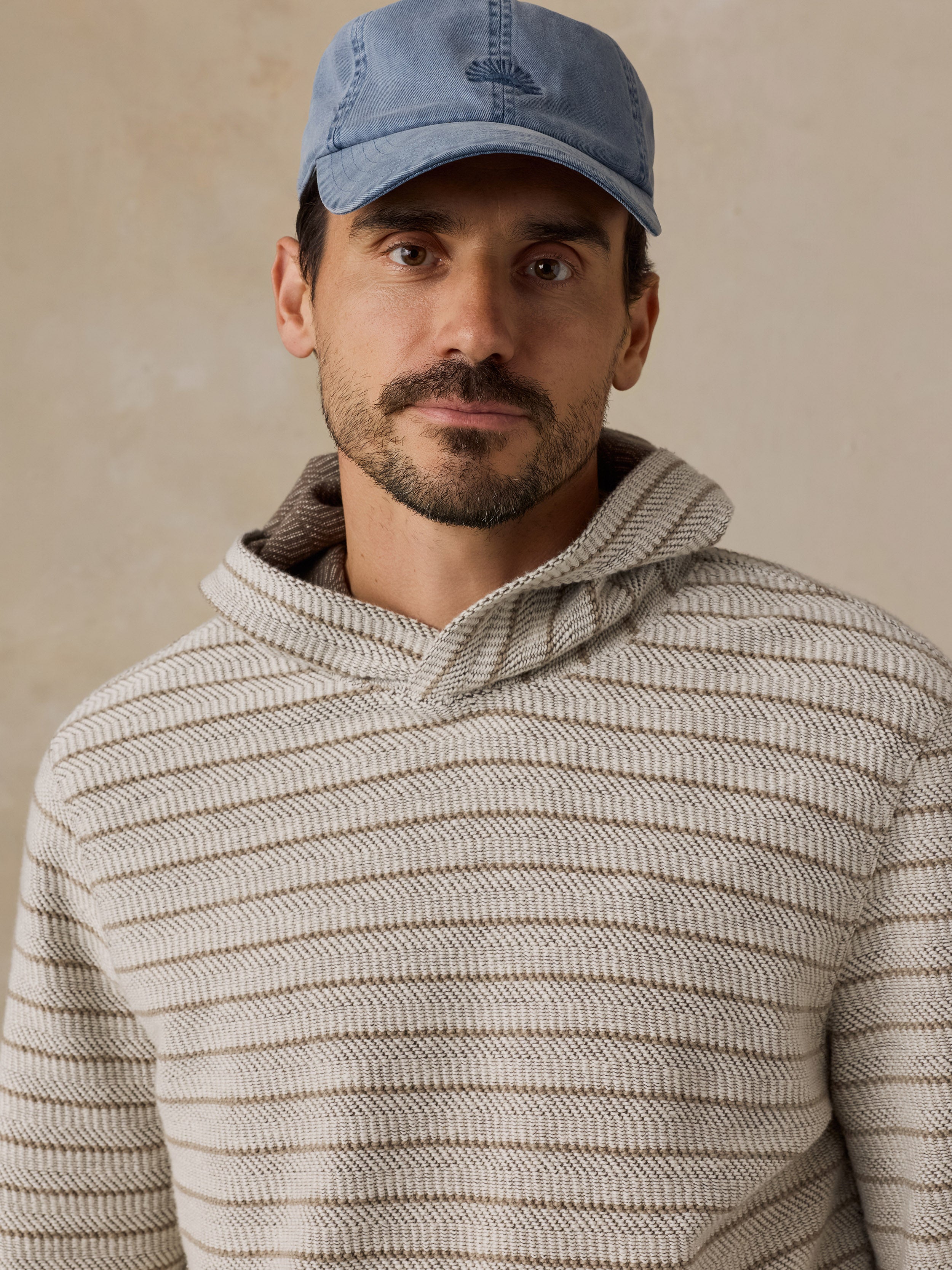 A man wearing a light blue baseball cap and a beige textured knit hoodie, photographed against a neutral beige background.