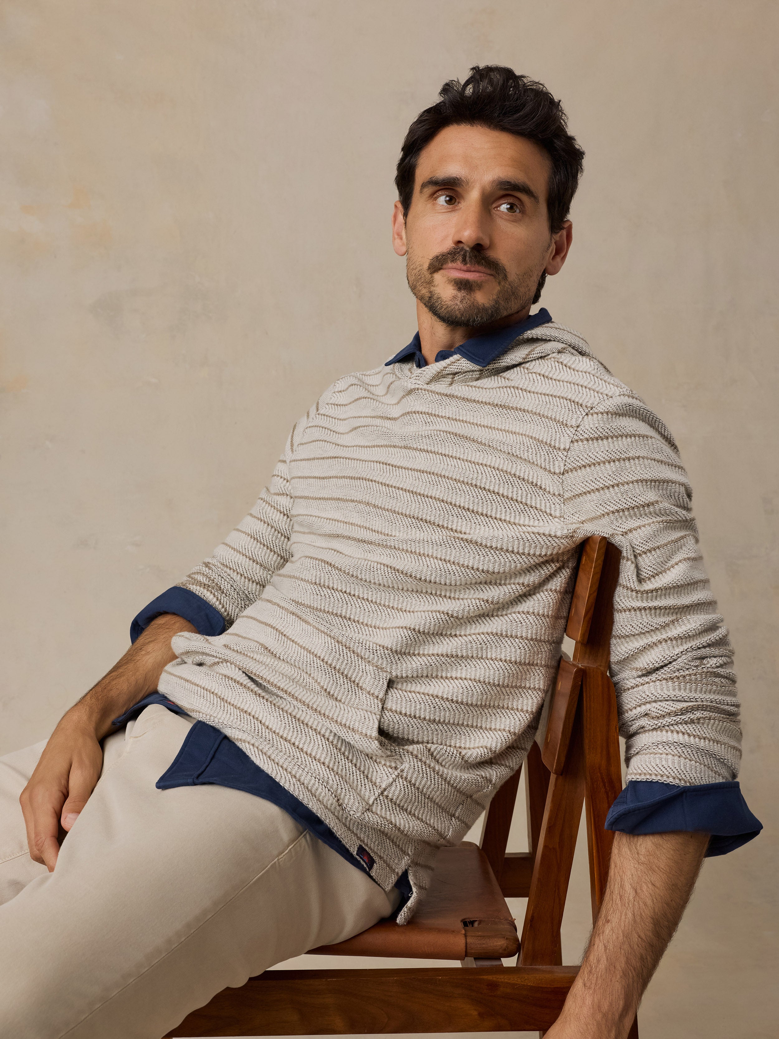 A man wearing a beige and white striped knit sweater over a blue collared shirt, paired with beige trousers, sitting on a wooden chair against a textured beige wall.