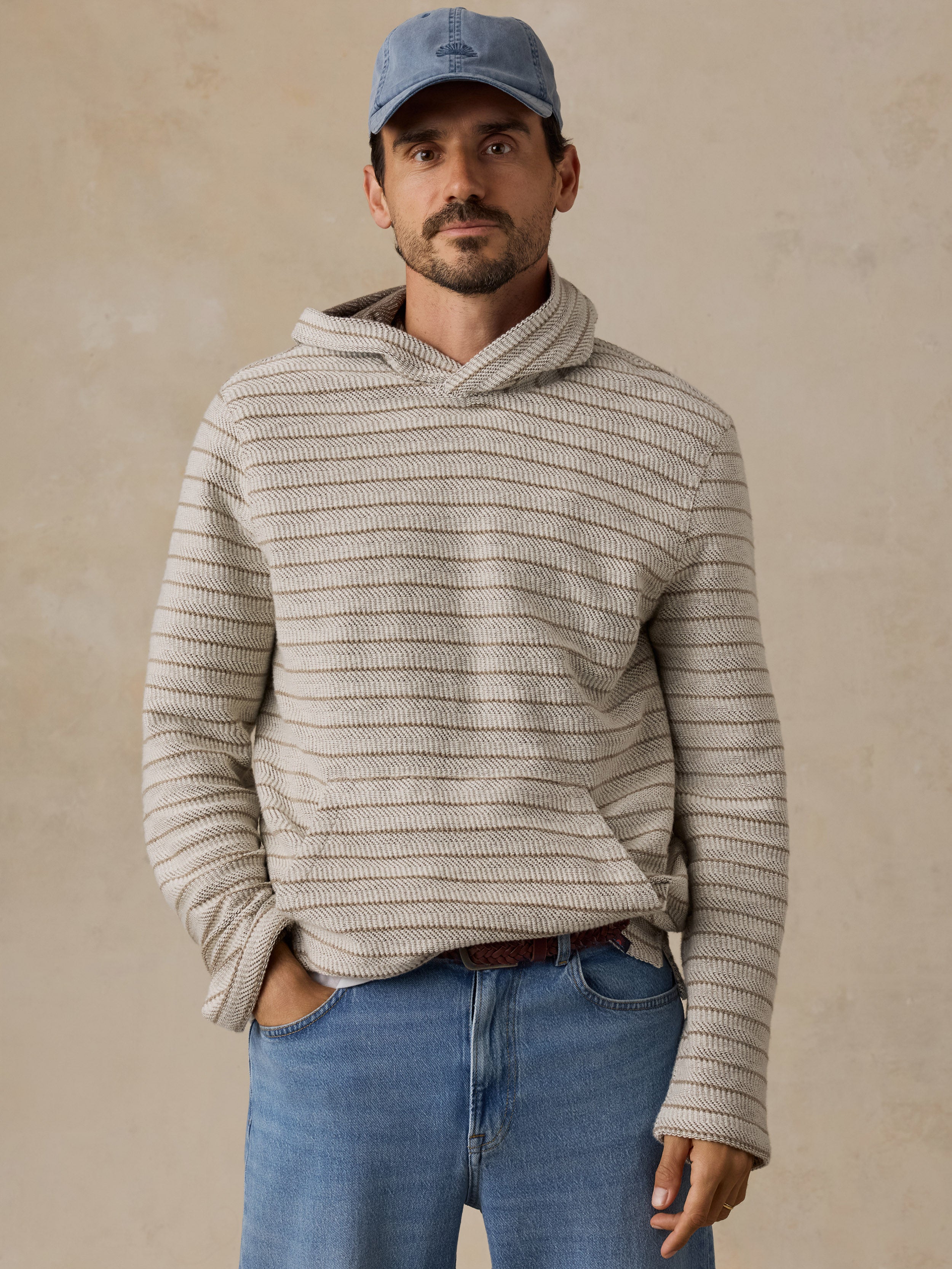 A man wearing a beige and gray horizontally striped knit hoodie with a cowl neck, paired with blue jeans and a blue baseball cap, standing against a textured beige wall.