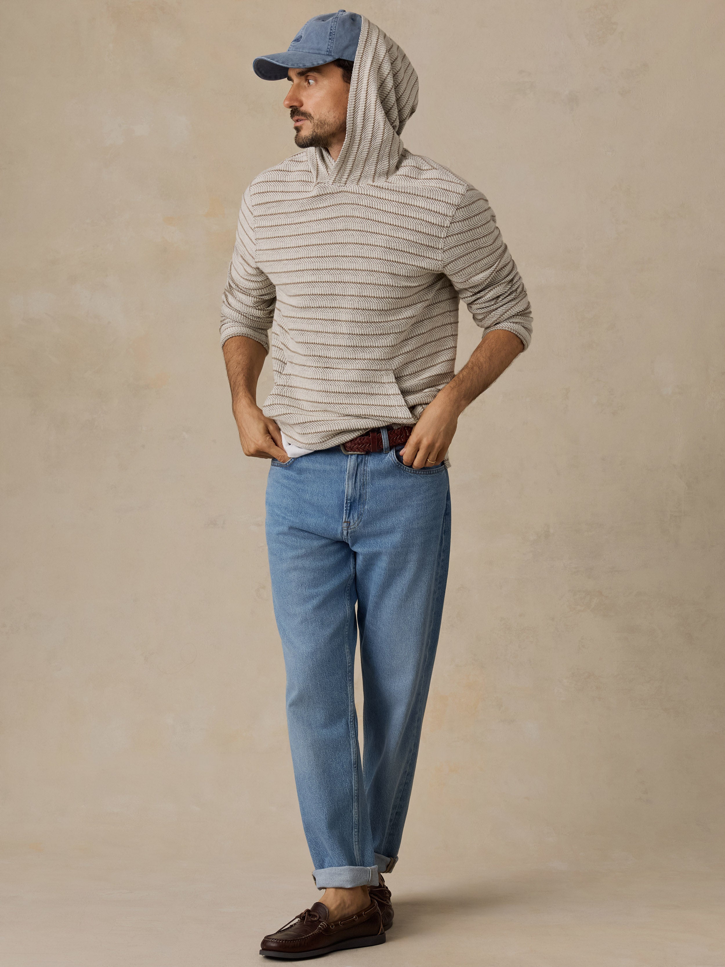 A man wearing a beige and gray striped hoodie, blue jeans, a blue baseball cap, and brown leather loafers, standing against a textured beige studio backdrop.