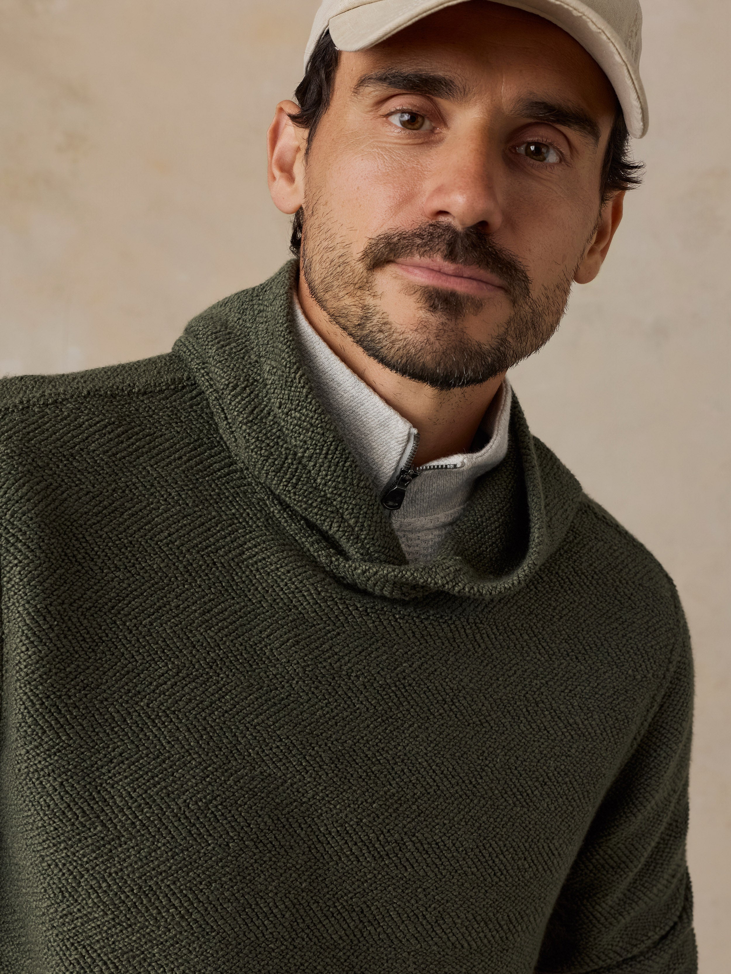 A close-up of a man wearing an olive green textured knit cowl-neck sweater over a light gray collared shirt, with a beige baseball cap, against a neutral beige background.