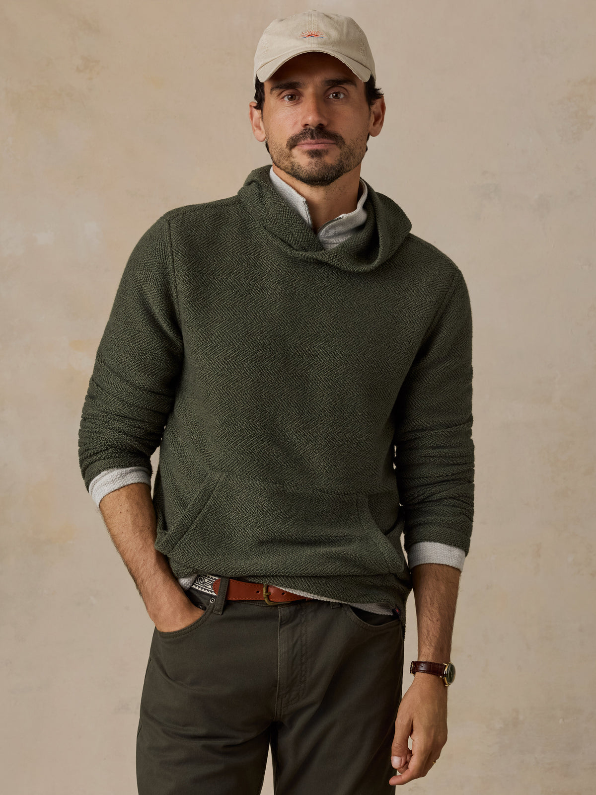 A man wearing an olive green cowl-neck knit sweater over a light gray collared shirt, paired with dark olive pants, a beige baseball cap, and a brown belt, standing against a textured beige wall.