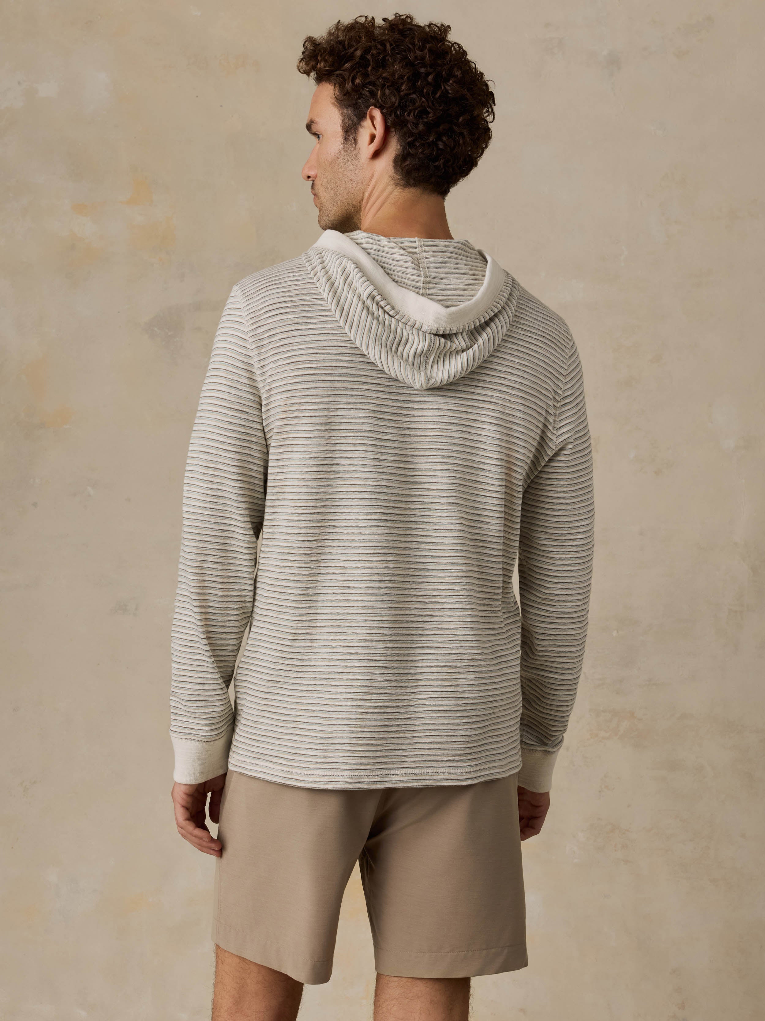 A man shown from the back wearing a light beige and gray striped hooded sweater and tan shorts, standing against a textured beige wall.