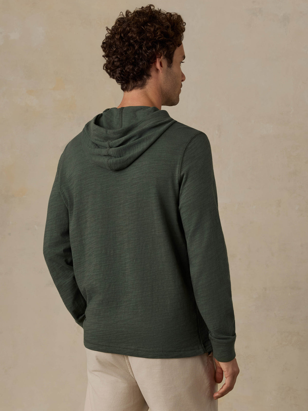 Man wearing a dark olive green hoodie and beige shorts against a beige background, image 3 of 6