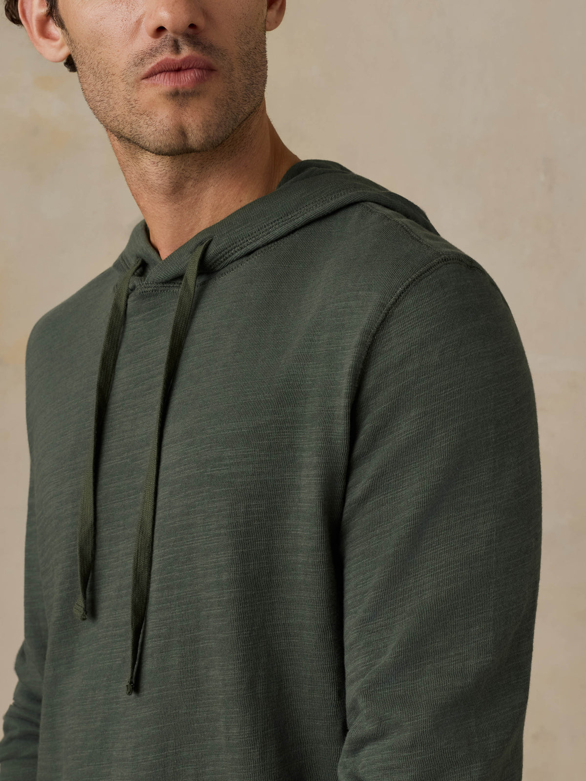 Close-up of a man wearing a dark olive green hoodie against a beige background, image 6 of 6