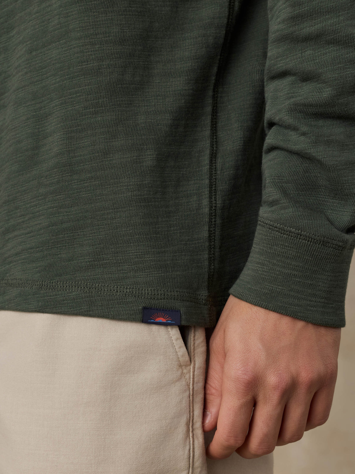 Close-up of a man wearing a dark olive green hoodie and beige shorts against a beige background, image 4 of 6