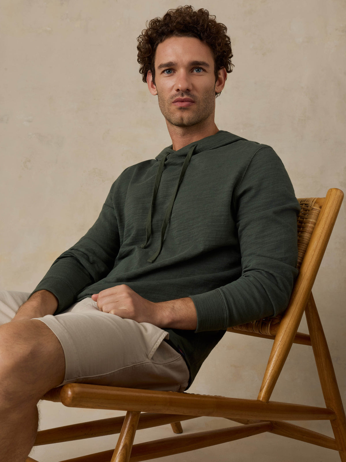 Man sitting on a wooden chair wearing a dark olive green hoodie and beige shorts against a beige background, image 2 of 6