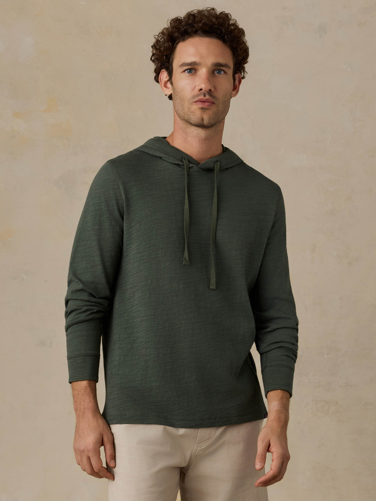 Man wearing a dark olive green hoodie and beige shorts against a beige background, image 1 of 6
