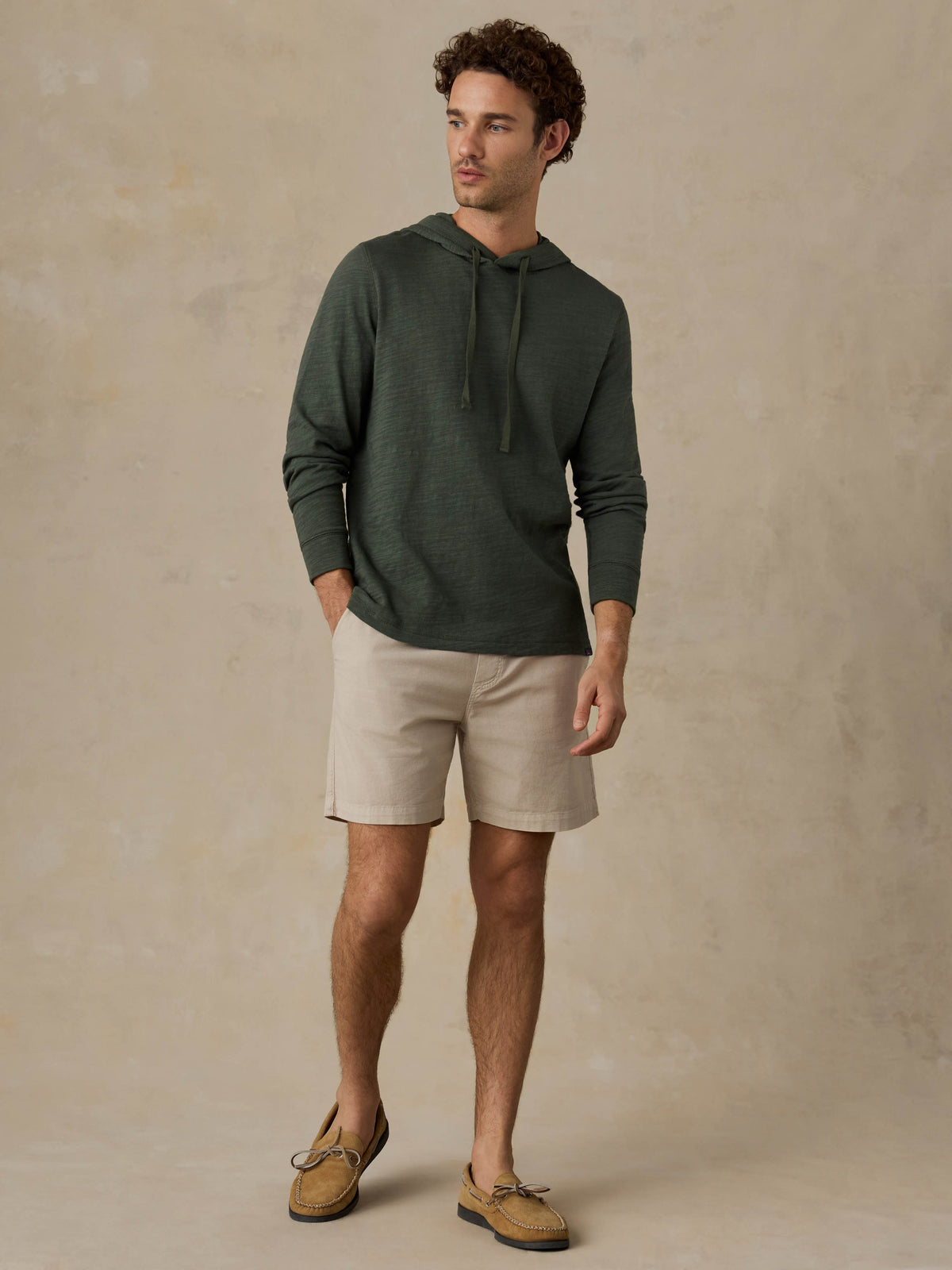 Man wearing a dark olive green hoodie, beige shorts, and boat shoes against a beige background, image 5 of 6