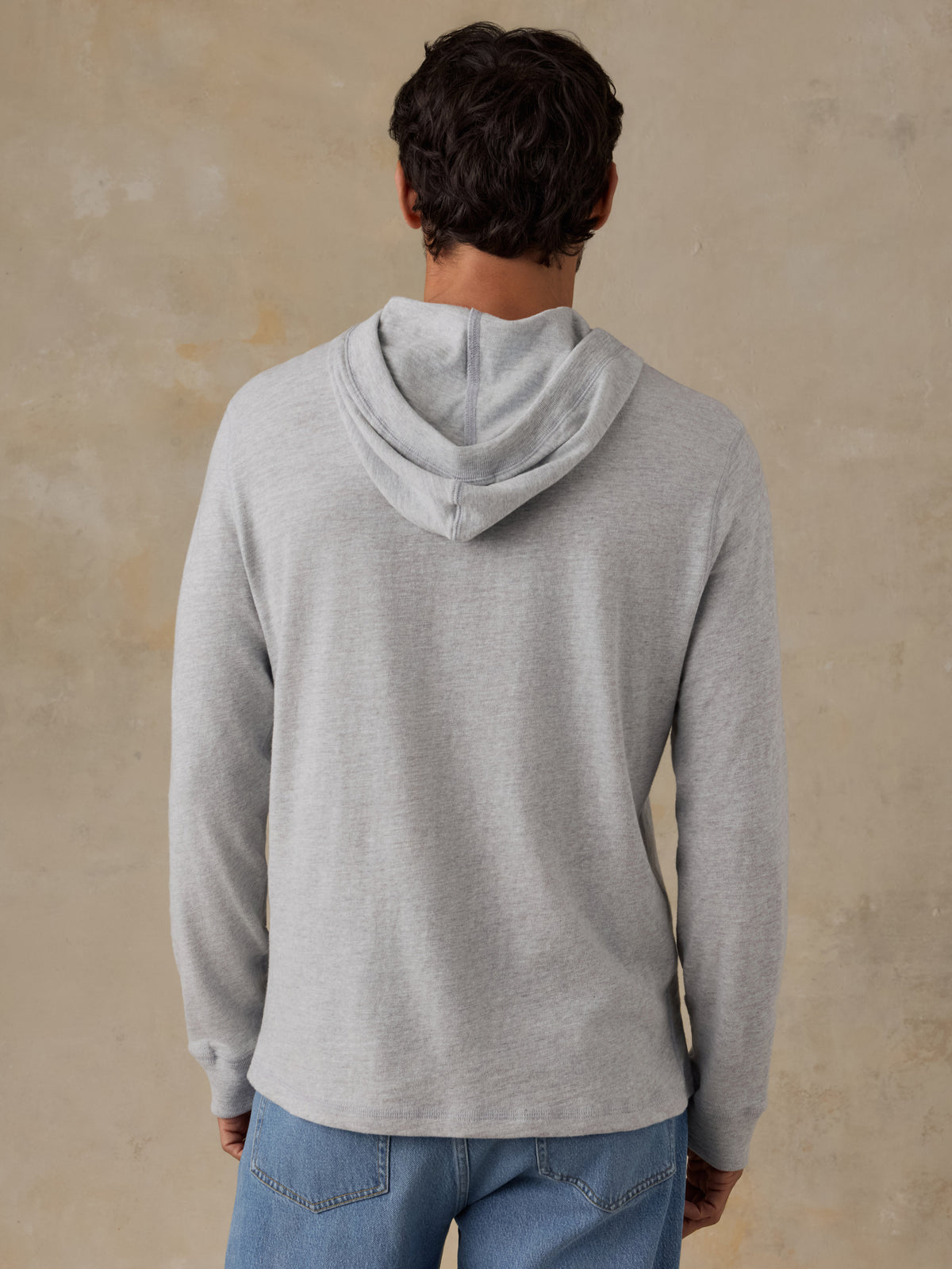 A man viewed from behind wearing a light gray heathered hooded sweatshirt and blue jeans, standing against a textured beige wall., image 3 of 8